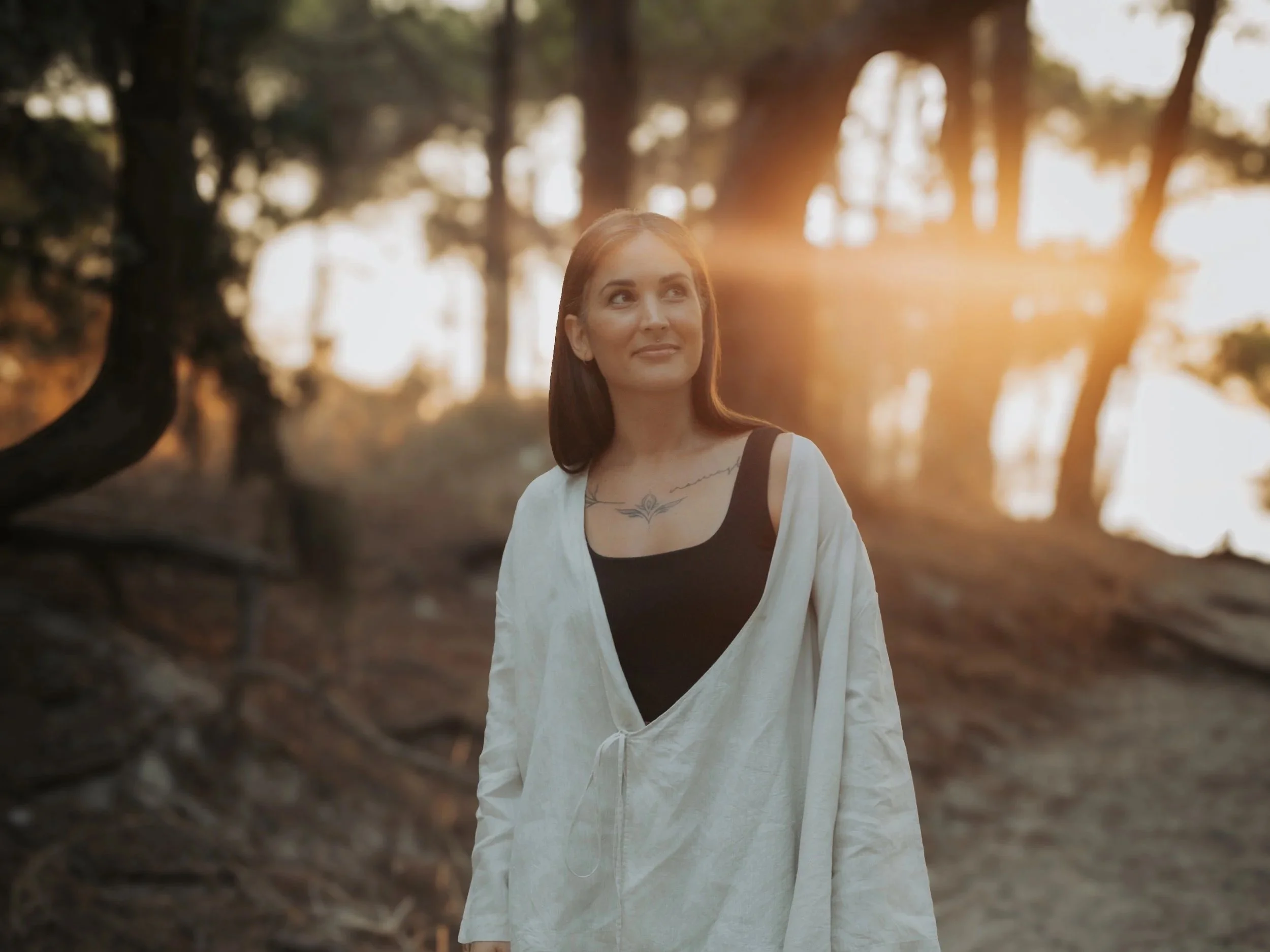 Una donna con capelli castani, indossa una gonna bianca e una maglietta nera, cammina nel bosco al tramonto con i raggi del sole che filtrano tra gli alberi.