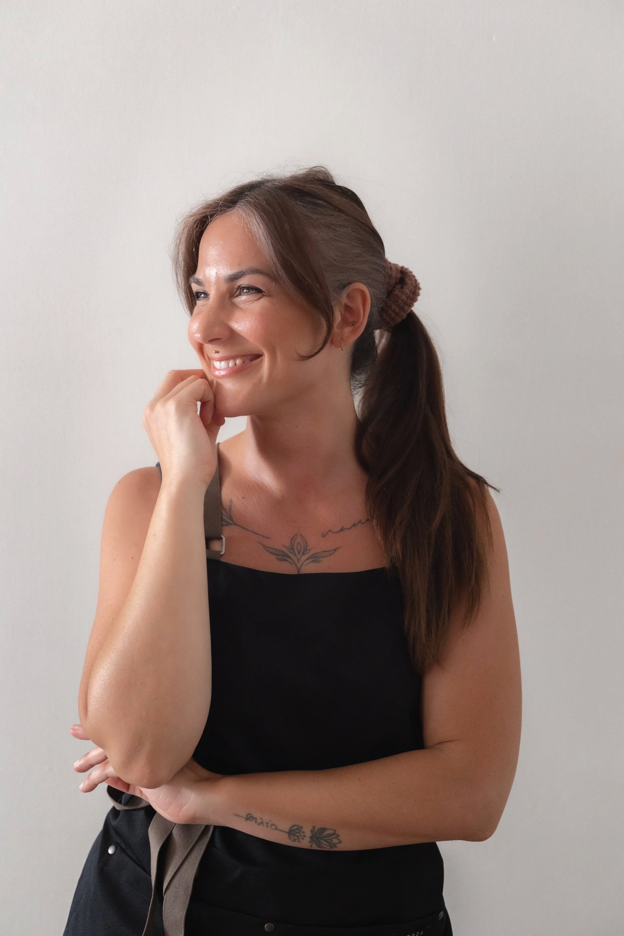 Una donna sorridente con i capelli raccolti in una coda, senza maglietta, con tatuaggi sul braccio e sul petto, che si appoggia al muro bianco.