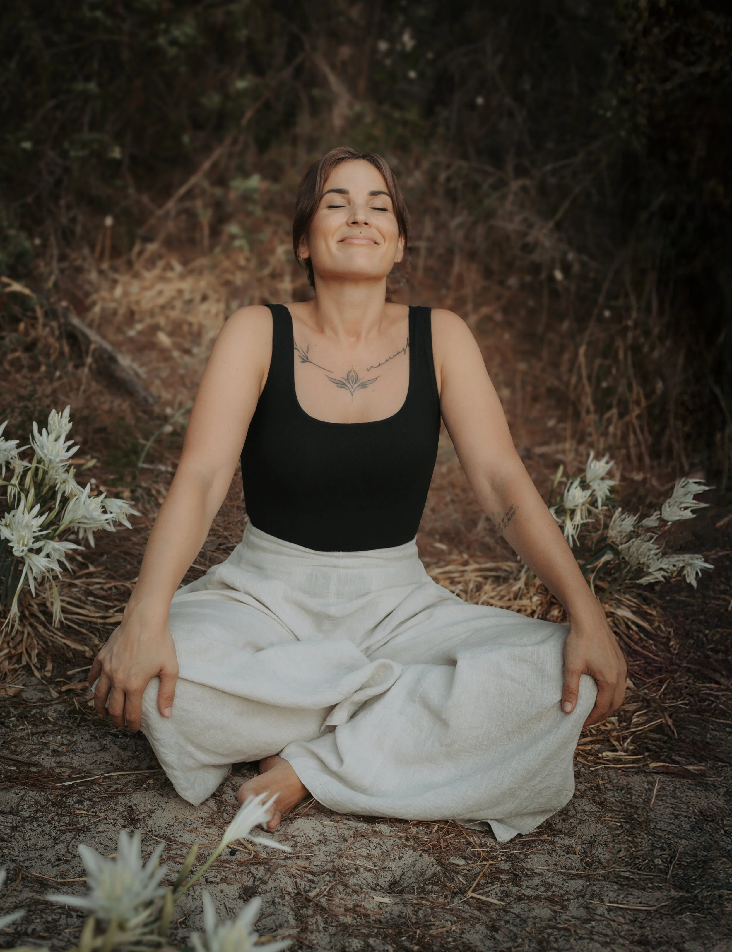 Una donna con capelli corti e tatuaggi sulla pelle, seduta a terra con gli occhi chiusi e un sorriso sereno, circondata da fiori bianchi in un ambiente naturale con erba e foglie.