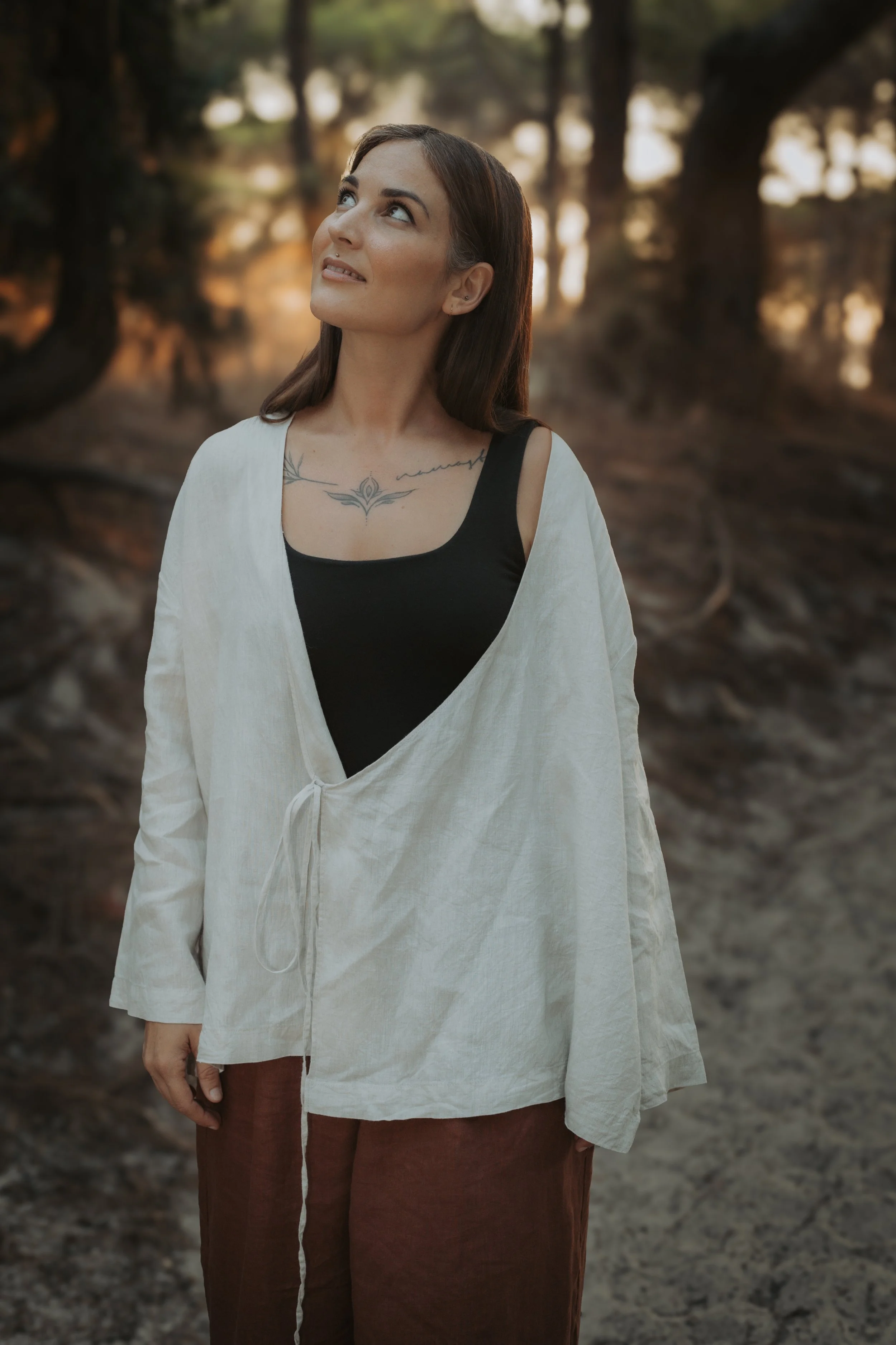 Donna con tatuaggio sul petto, vestita con una maglietta nera a maniche corte e una tunica bianca, in piedi in una foresta durante il tramonto