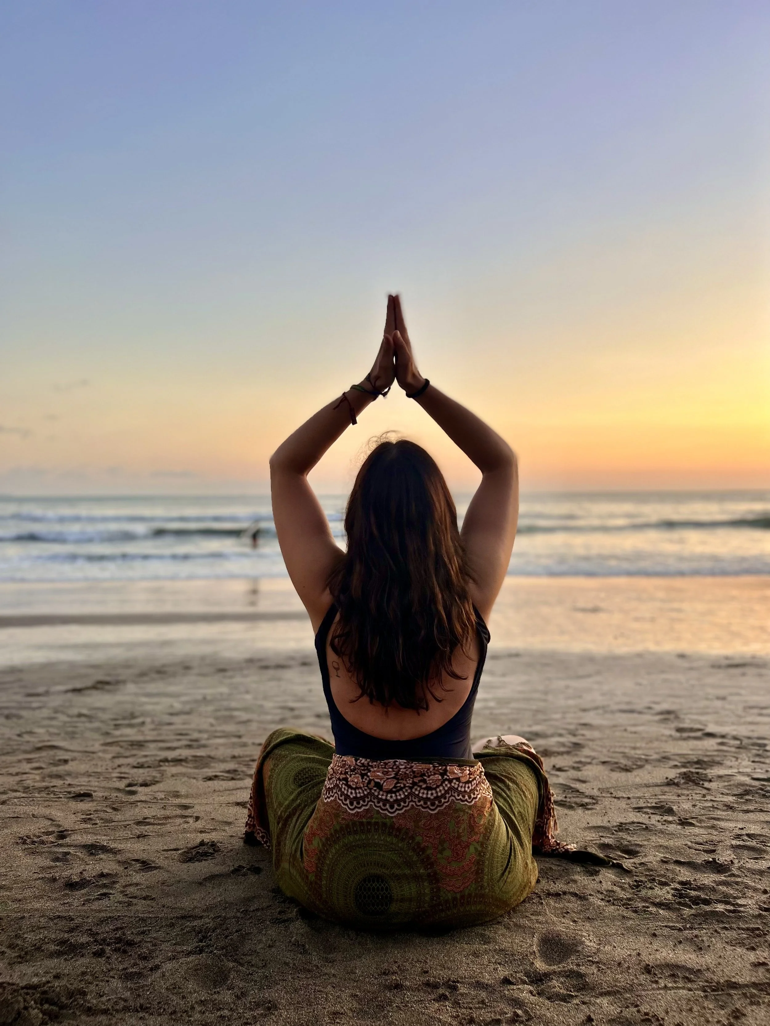 Donna seduta sulla spiaggia che fa yoga con le gambe incrociate e le mani giunte sopra la testa durante il tramonto.