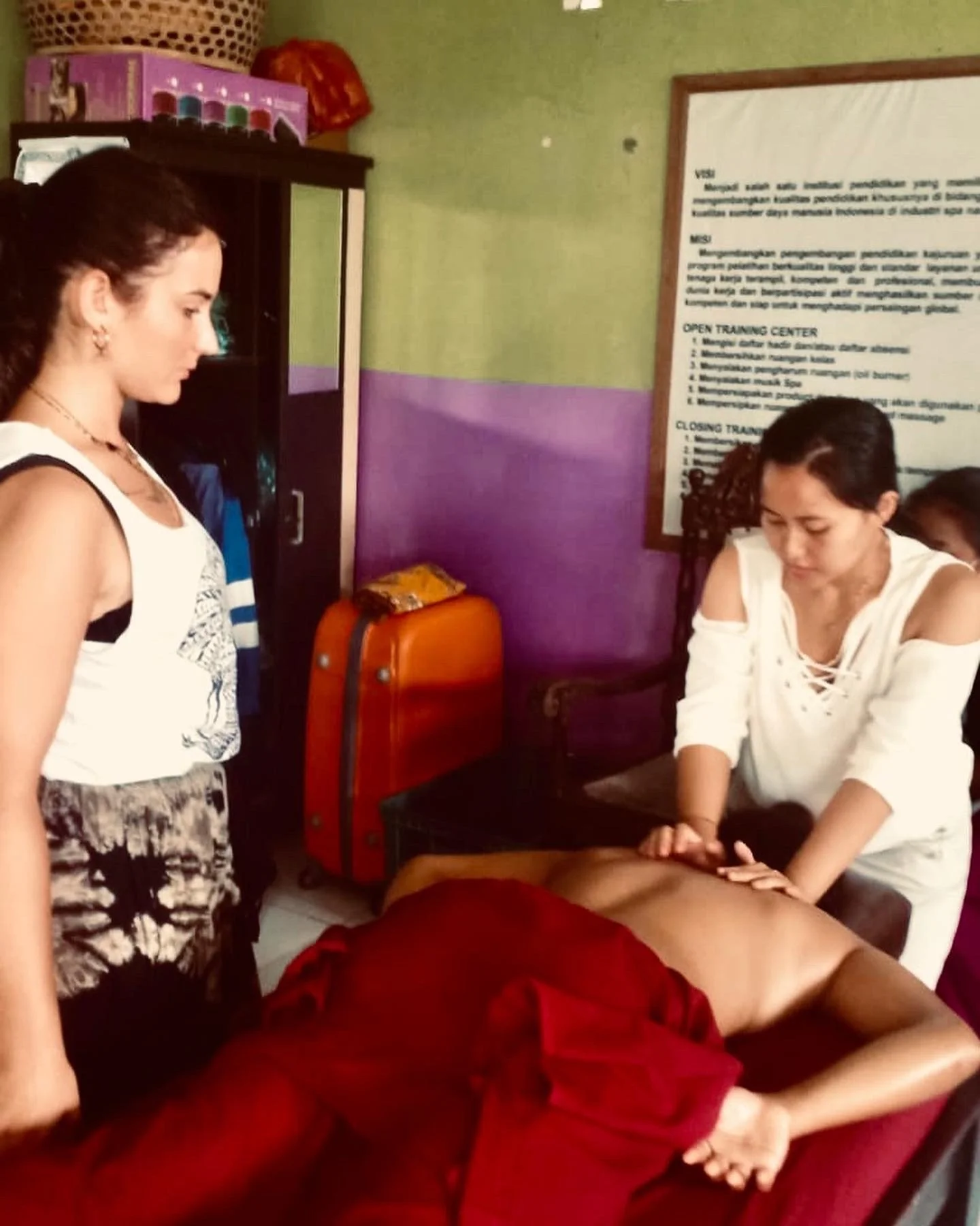 Due donne stanno praticando un'acupressione su un uomo sdraiato, in un ambiente di tipo clinico o di formazione, con un cartello informativo sullo sfondo.