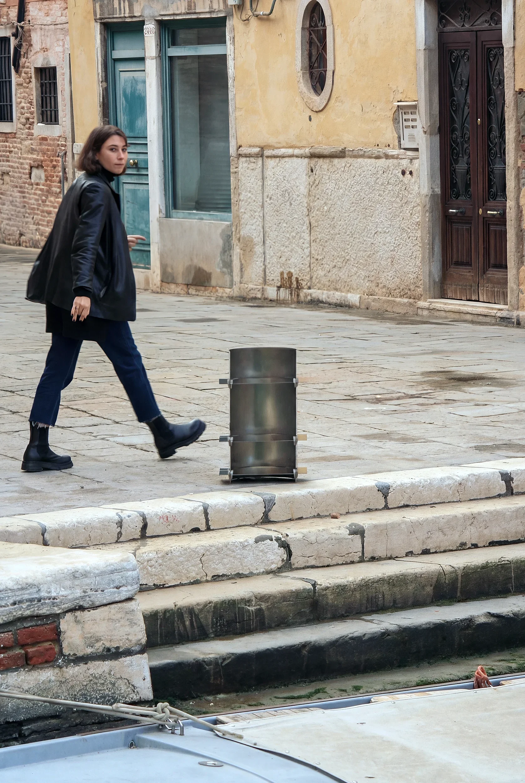 Una donna con una giacca nera e pantaloni blu cammina sulla strada accanto a una banchina di pietra, con un edificio antico sullo sfondo e un contenitore metallico accanto a lei.
