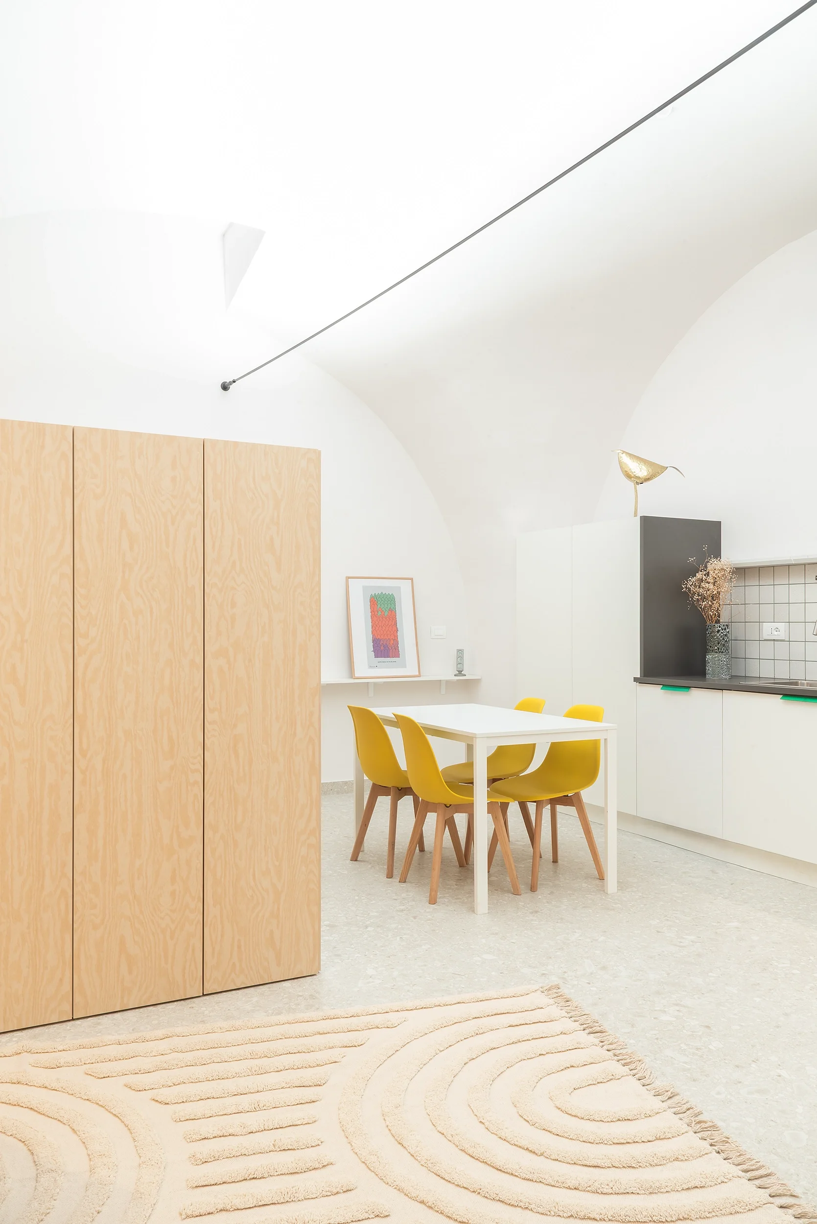 Interno di una cucina moderna e minimalista con pareti bianche, tavolo bianco con sedie gialle, armadio in legno chiaro, e decorazioni come un quadro, una pianta e una scultura a forma di uccello