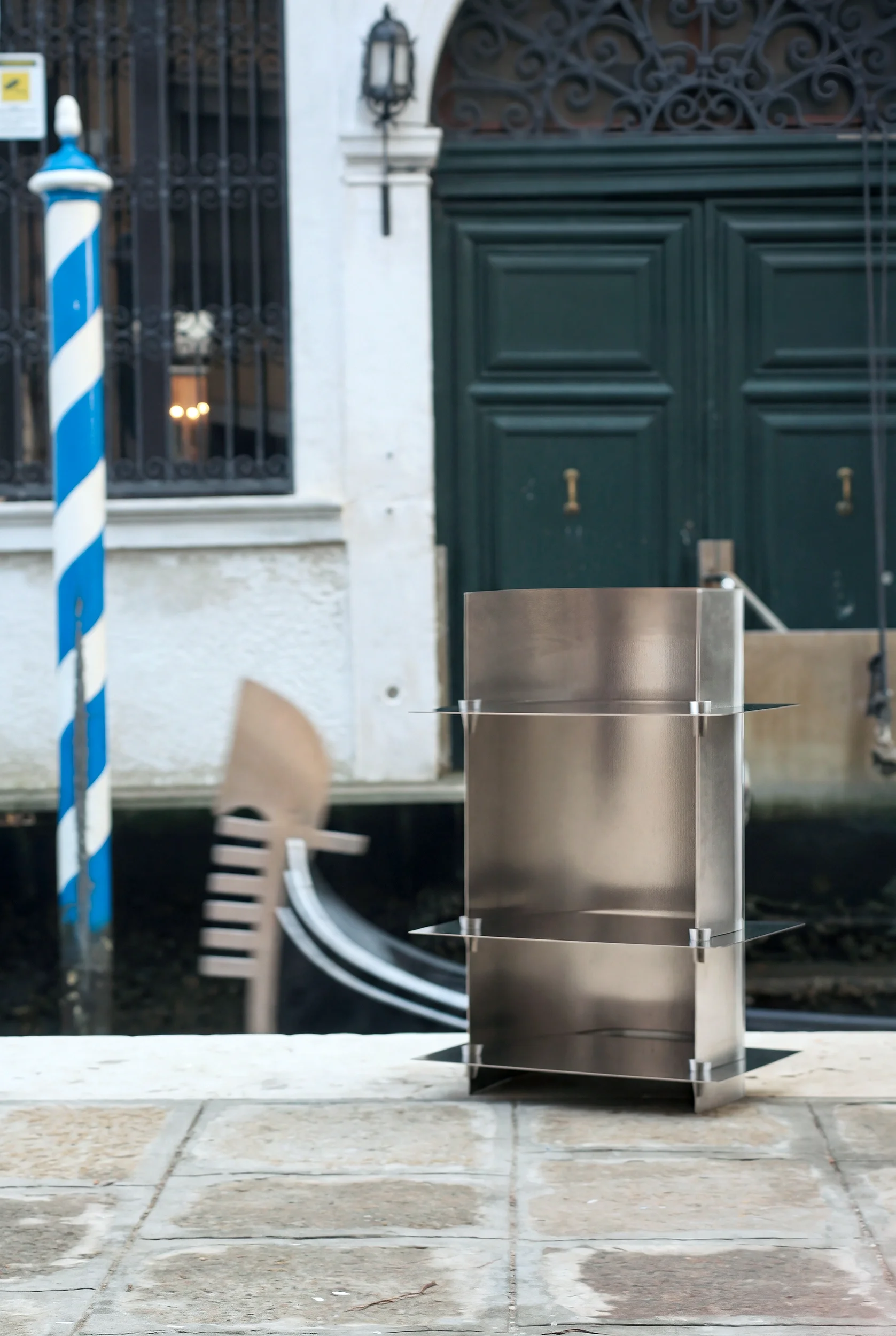 Banca di metallo in un'area all'aperto di fronte a un cancello verde e una parete bianca, con elementi decorativi come un lampione e un palo con strisce blu e bianche.