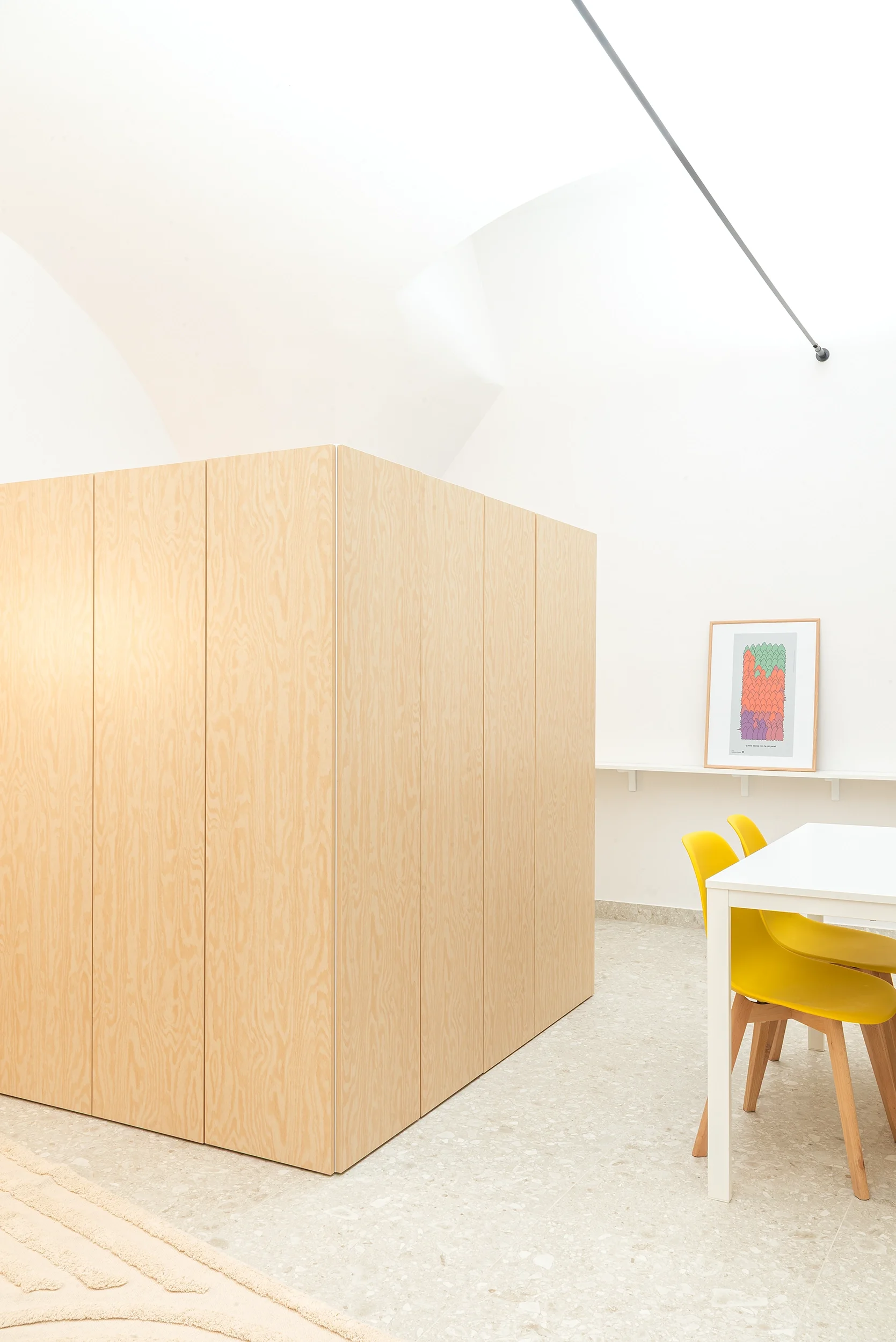 Interno minimalista con mobile in legno chiaro, tavolo bianco, sedie gialle e quadro astratto colorato alla parete.