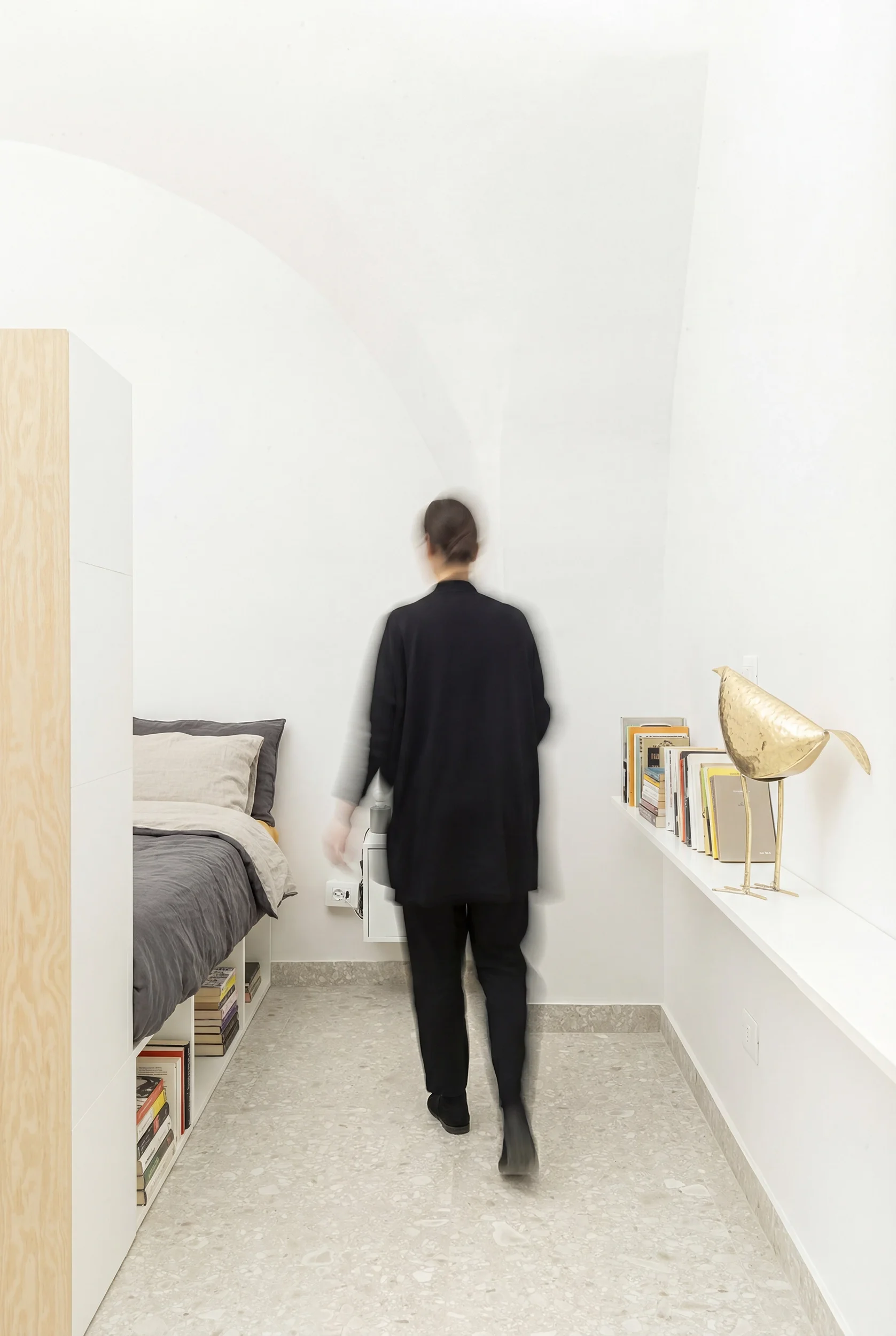 Una persona vestita di nero cammina in una stanza minimalista con pareti bianche, libri su una mensola e una decorazione a forma di uccello dorato.