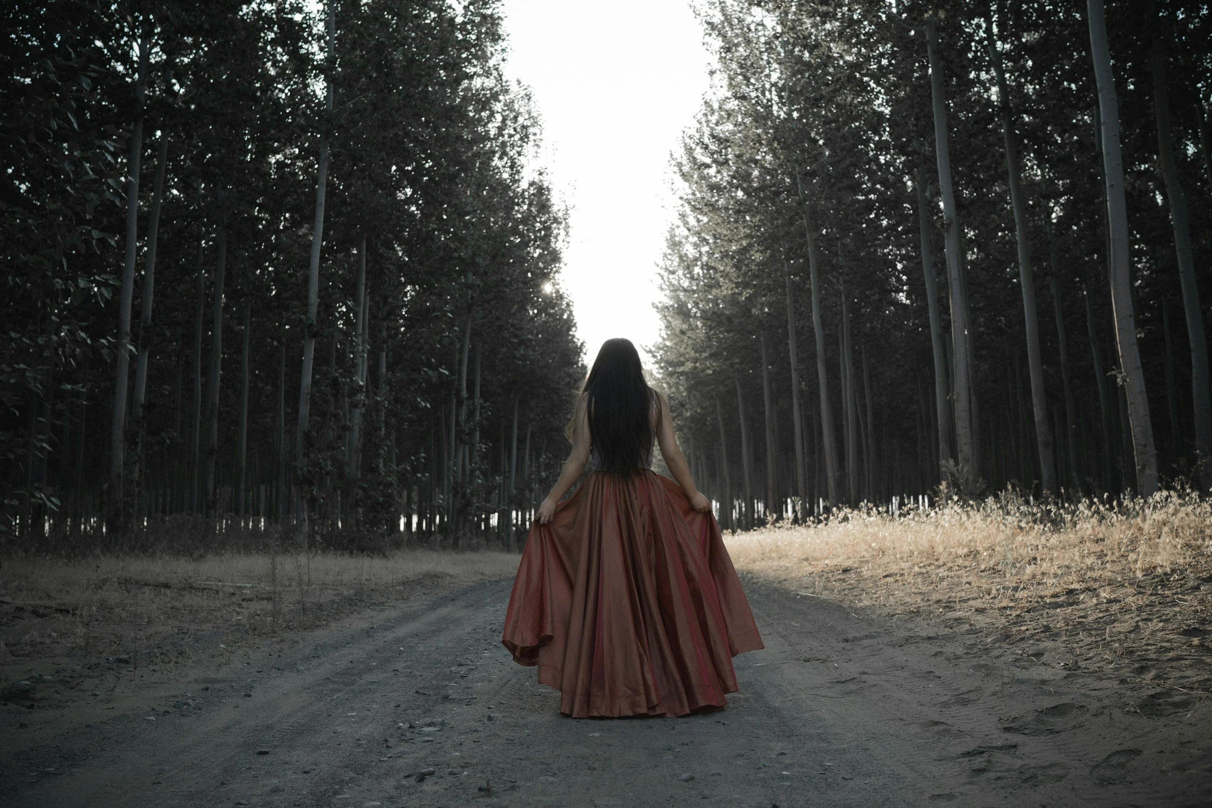 Woman in a rust-colored skirt walking through the forest at sunset, representing feminine healing, identity reclamation, and emotional renewal.