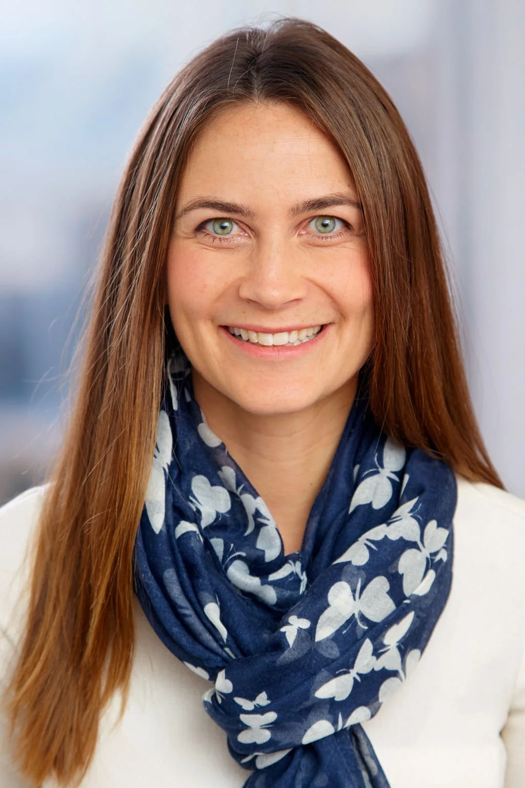 A woman with long brown hair, blue eyes, and a bright smile, wearing a white top and a blue floral scarf, posed in front of a blurred outdoor background.