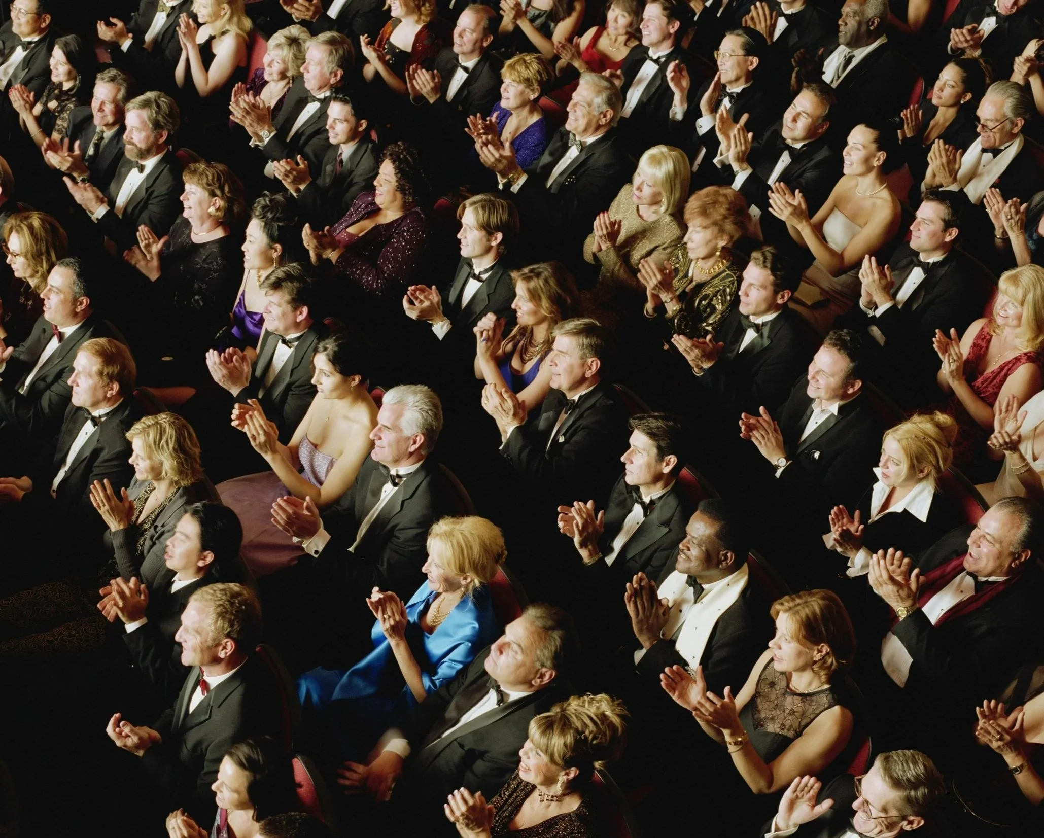 Audience members at a formal event, dressed in tuxedos and evening gowns, clapping and smiling, seated in rows inside a theater or auditorium.