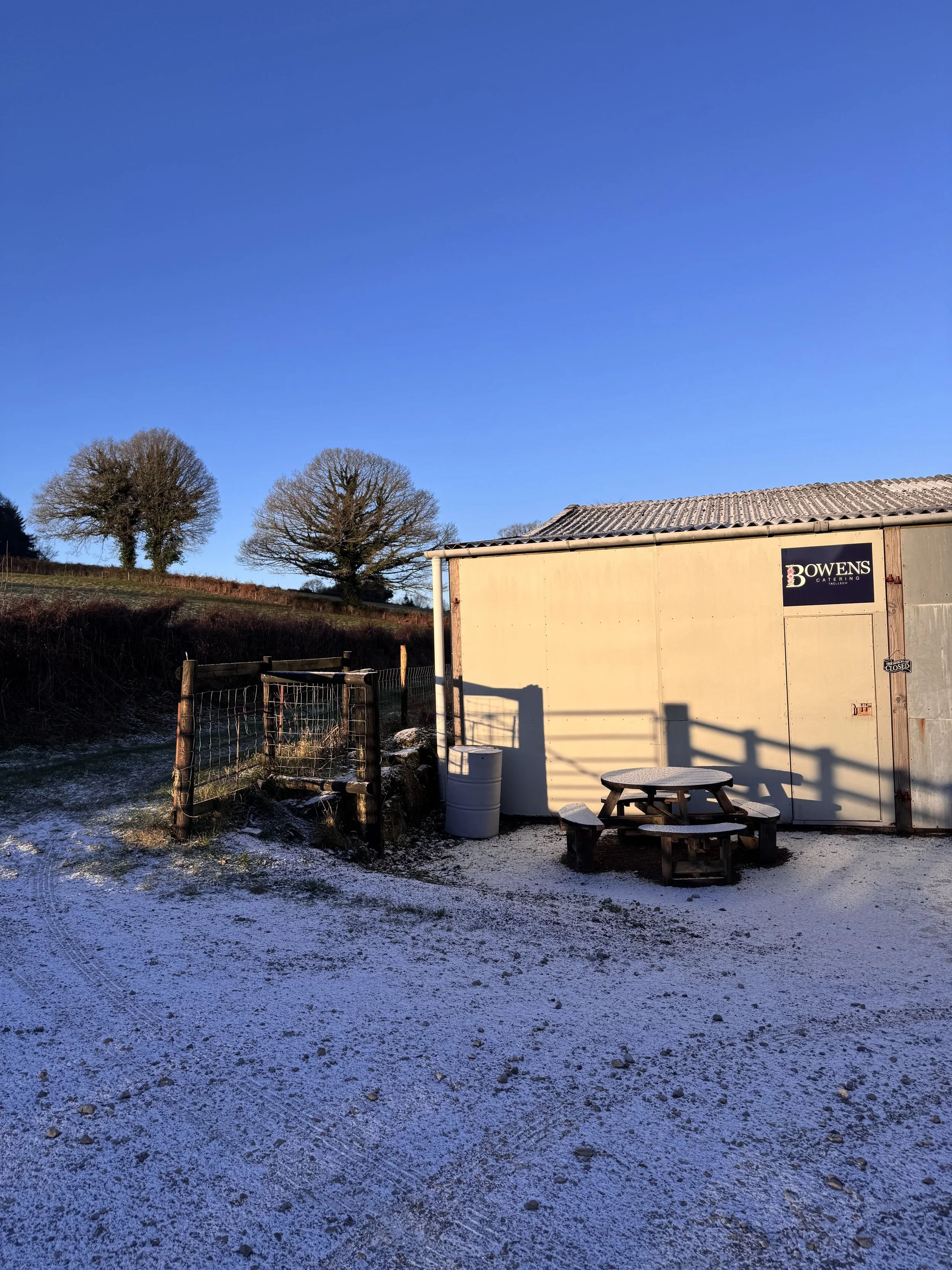 Sunlit outdoor scene with a small shed bearing a 'Bowens Catering' sign, a picnic table, a trash bin, a wooden fence, leafless trees on a grassy hill, and a clear blue sky.