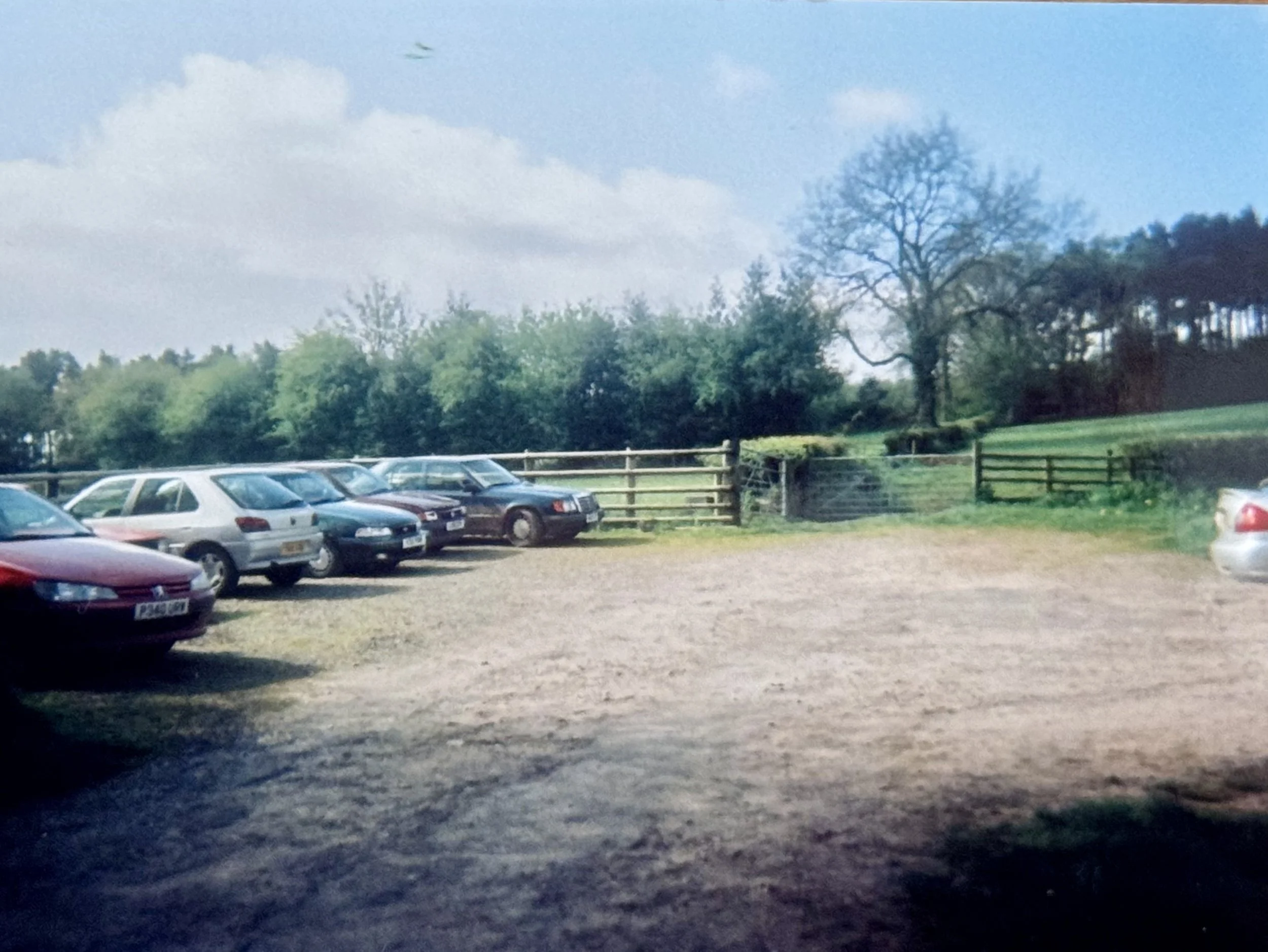 A rural parking lot with several parked cars, a wooden fence, green fields, and trees under a partly cloudy sky.