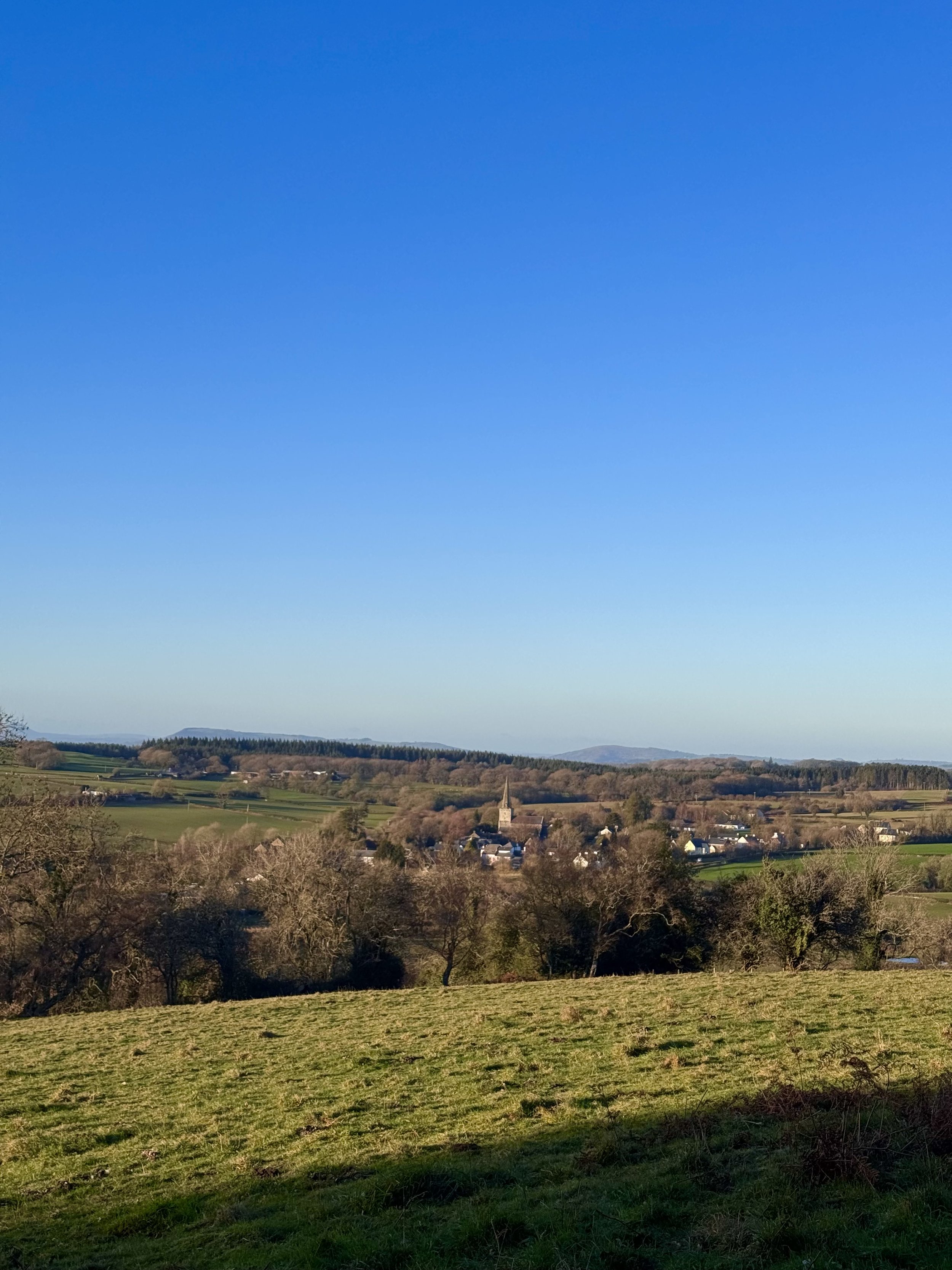 A scenic view of rolling green hills, a small village with white houses and a church steeple, and a clear blue sky in the background.