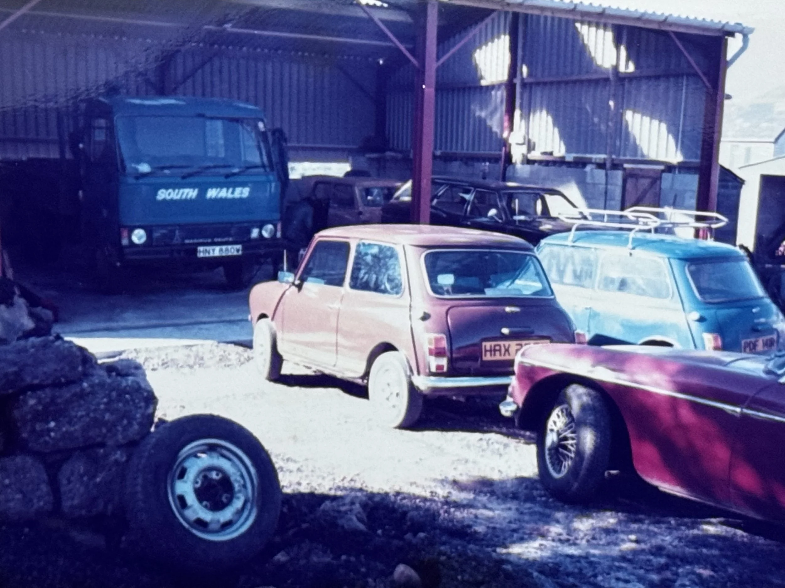 A vintage car garage with several classic cars parked inside, including a pink and purple car, a blue camper van, and other vehicles stored in a rustic structure with a corrugated metal roof.