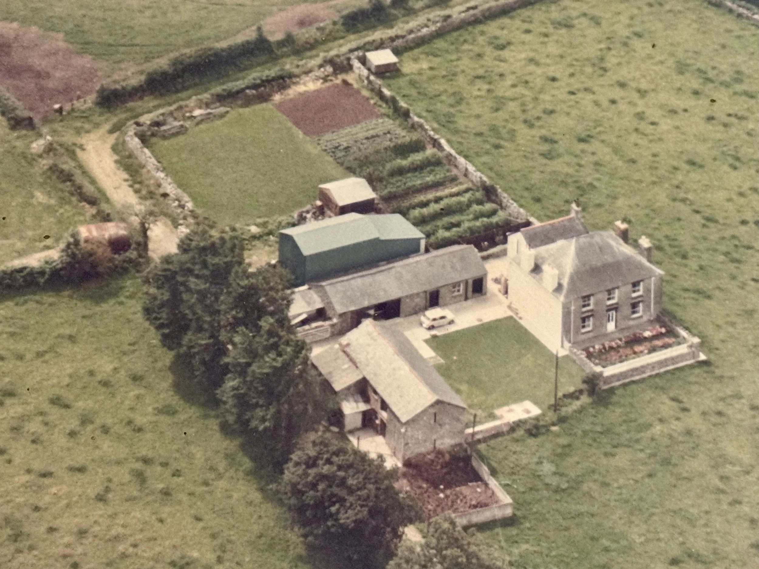 An aerial view of a rural farmstead with a main two-story house, a smaller outbuilding, a green barn, and a garden, surrounded by fields and patches of trees.