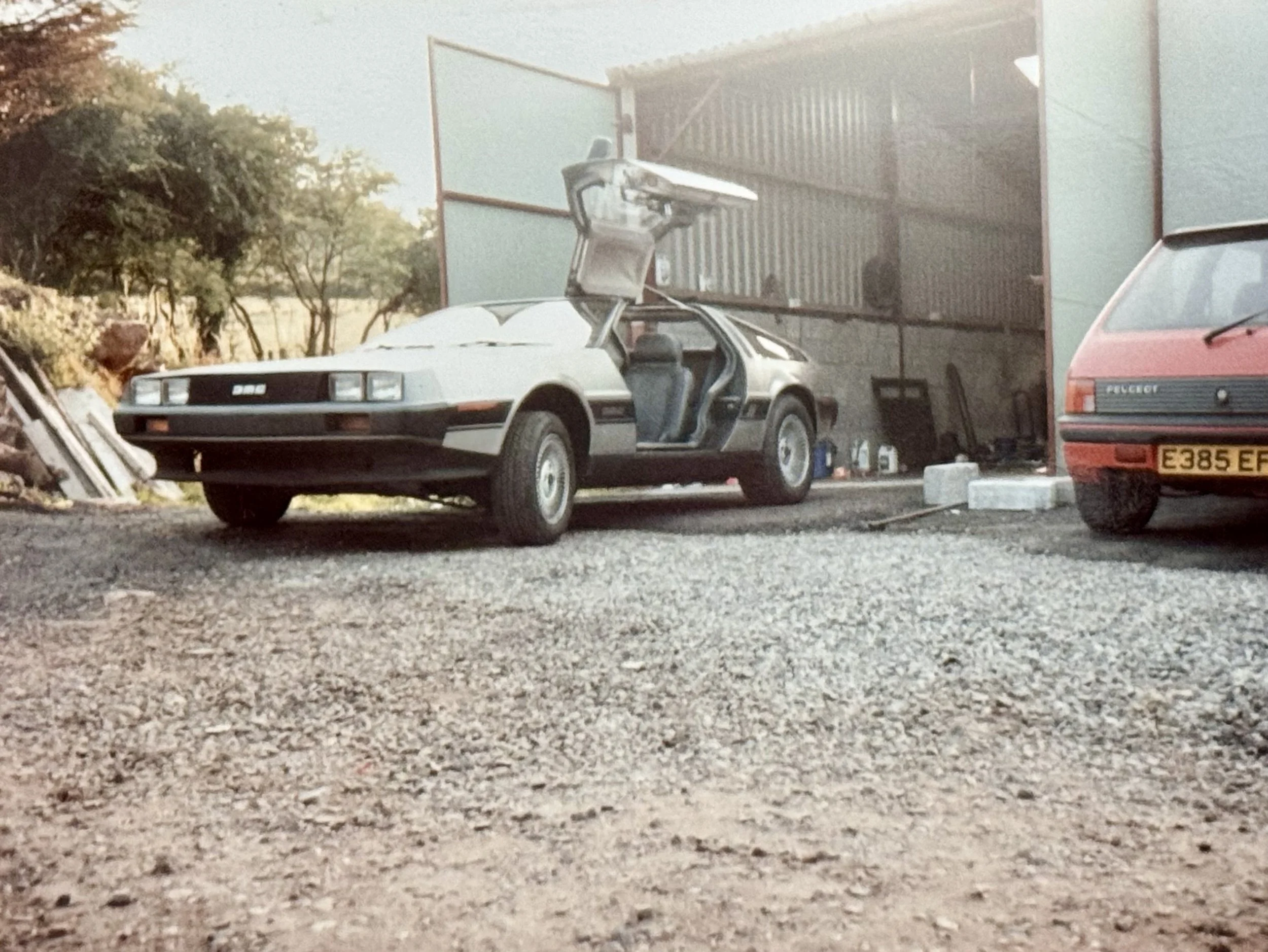 A classic silver DeLorean car with gull-wing doors open, parked outside a building with a red Peugeot car nearby.