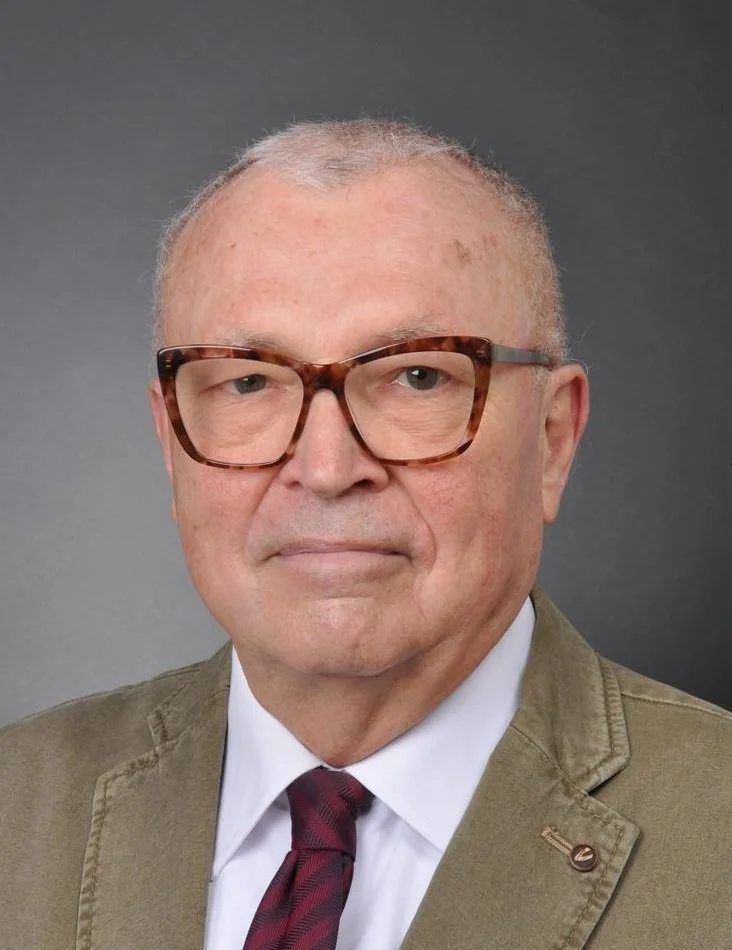 A middle-aged man with short gray hair wearing tortoise-shell glasses, a white shirt, a red and black striped tie, and a tan blazer against a gray background.