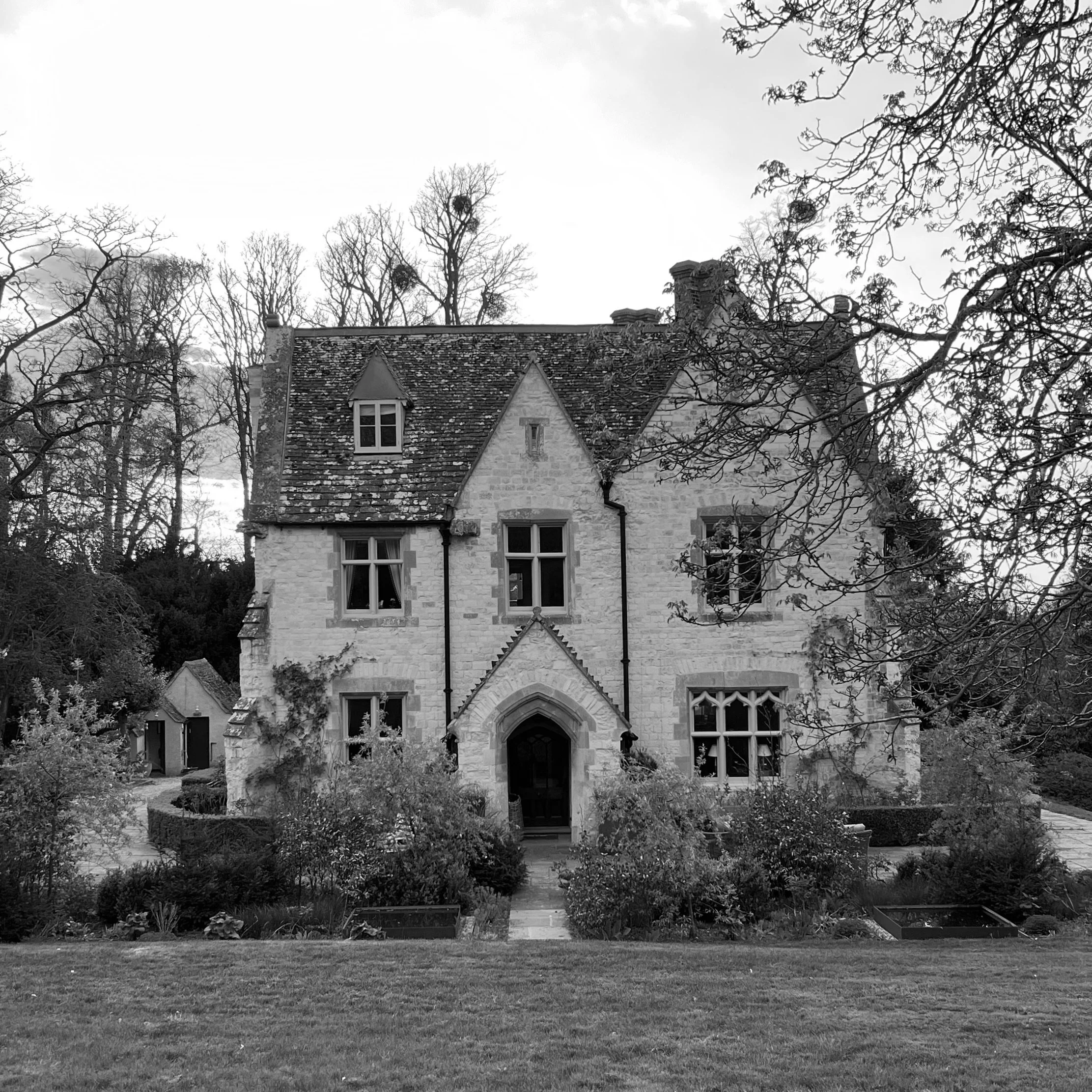 A historic stone house with a steep pitched roof, multiple windows, and surrounding trees in black and white.