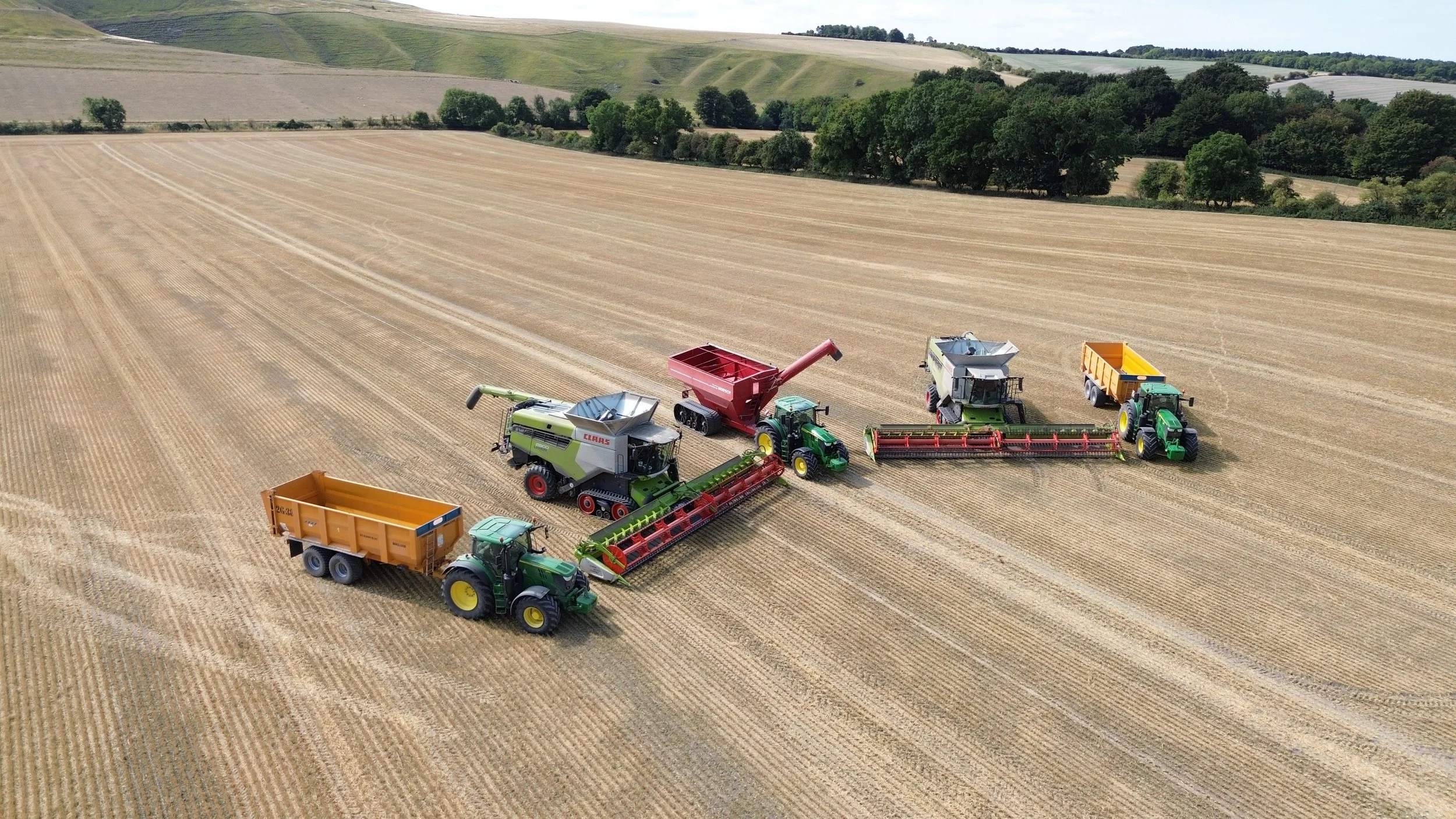Aerial view of multiple green tractors with various harvesting attachments working on a large wheat field with rolling hills and trees in the background.