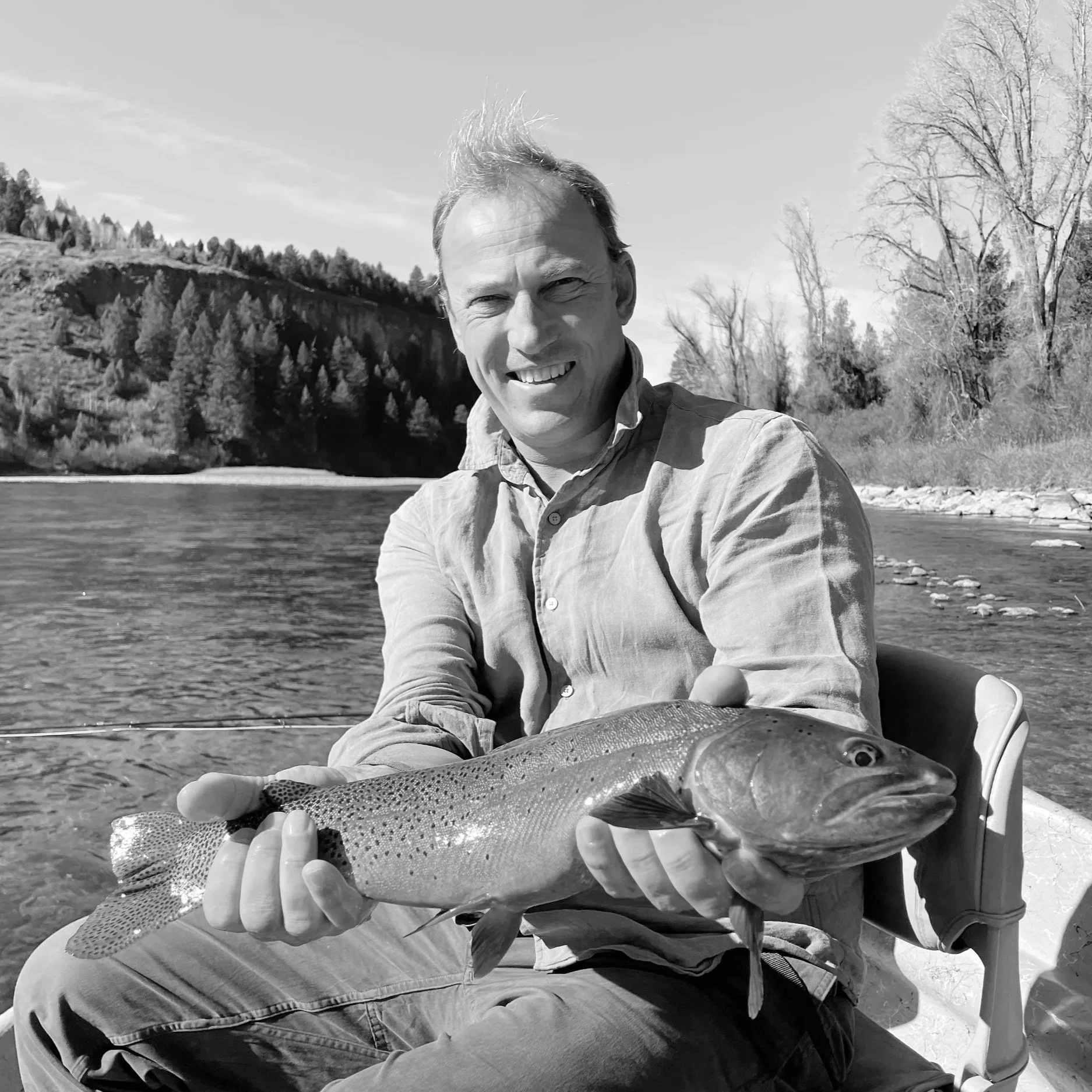 A man sitting in a boat holding a large fish, outdoors by a river with trees and a hillside in the background.
