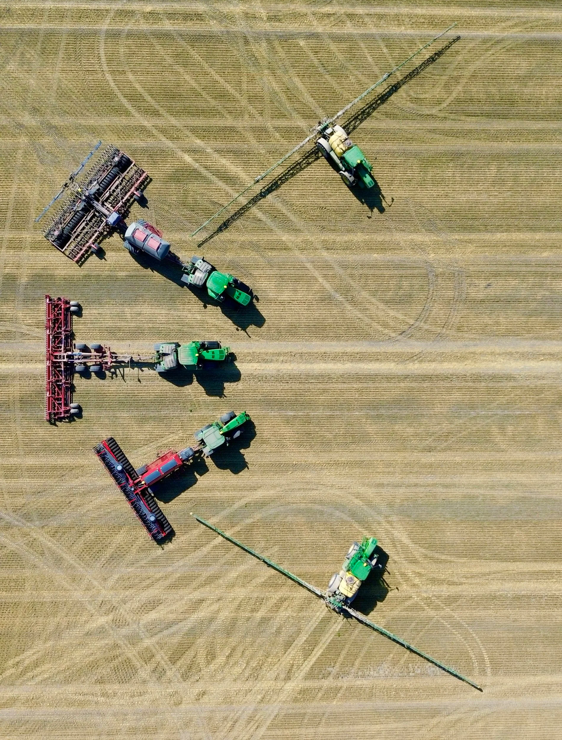 An aerial view of multiple green tractors and red farming equipment working in a large, golden wheat field.