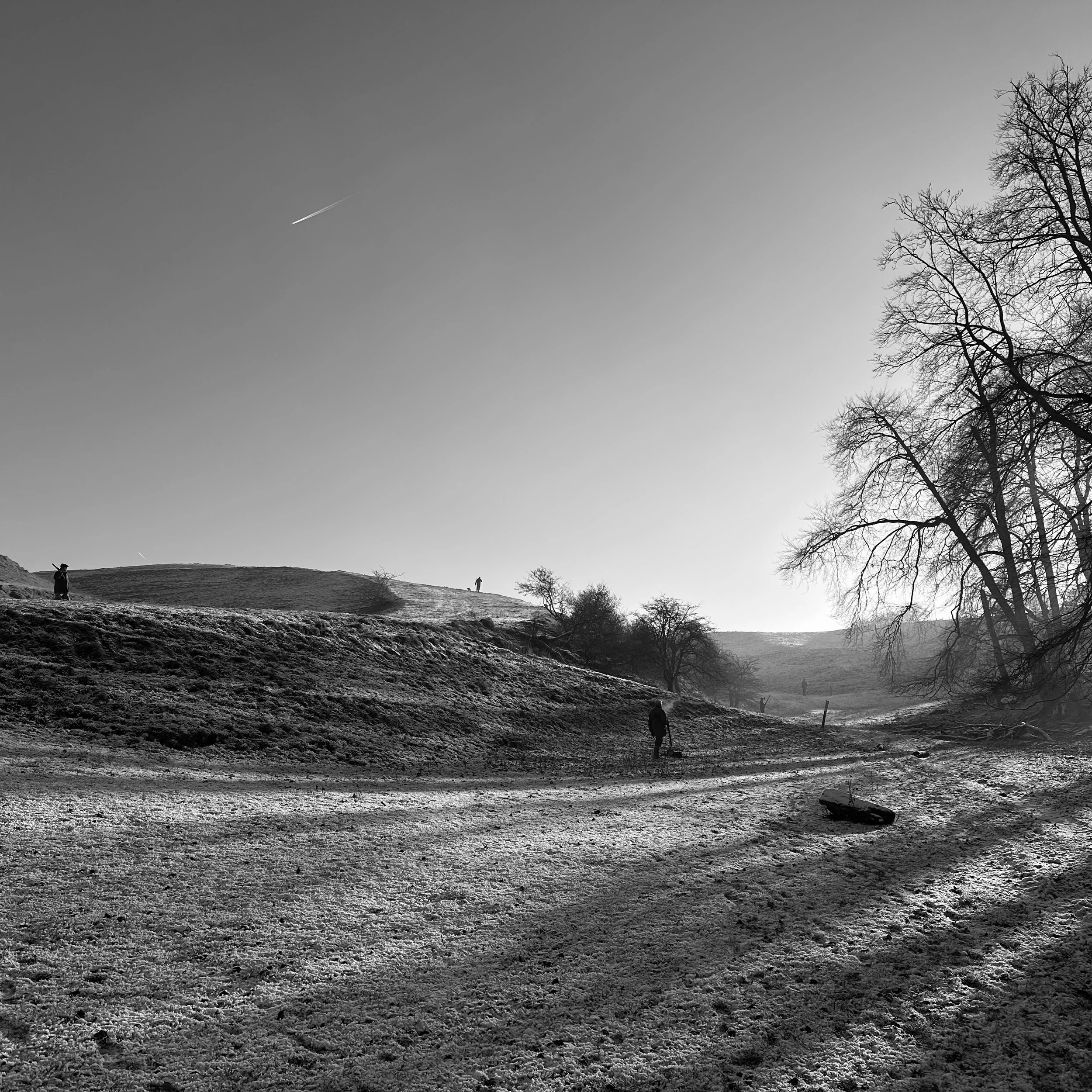 A black and white photo of a landscape with a dirt path, trees, and people walking or standing on the hills, with a clear sky with a contrail.