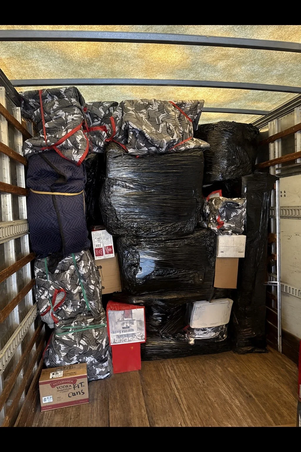 The image shows the inside of a truck cargo area filled with large black garbage bags, some with red and green ties, and several cardboard boxes, including one labeled 'Vodka' at the bottom left.