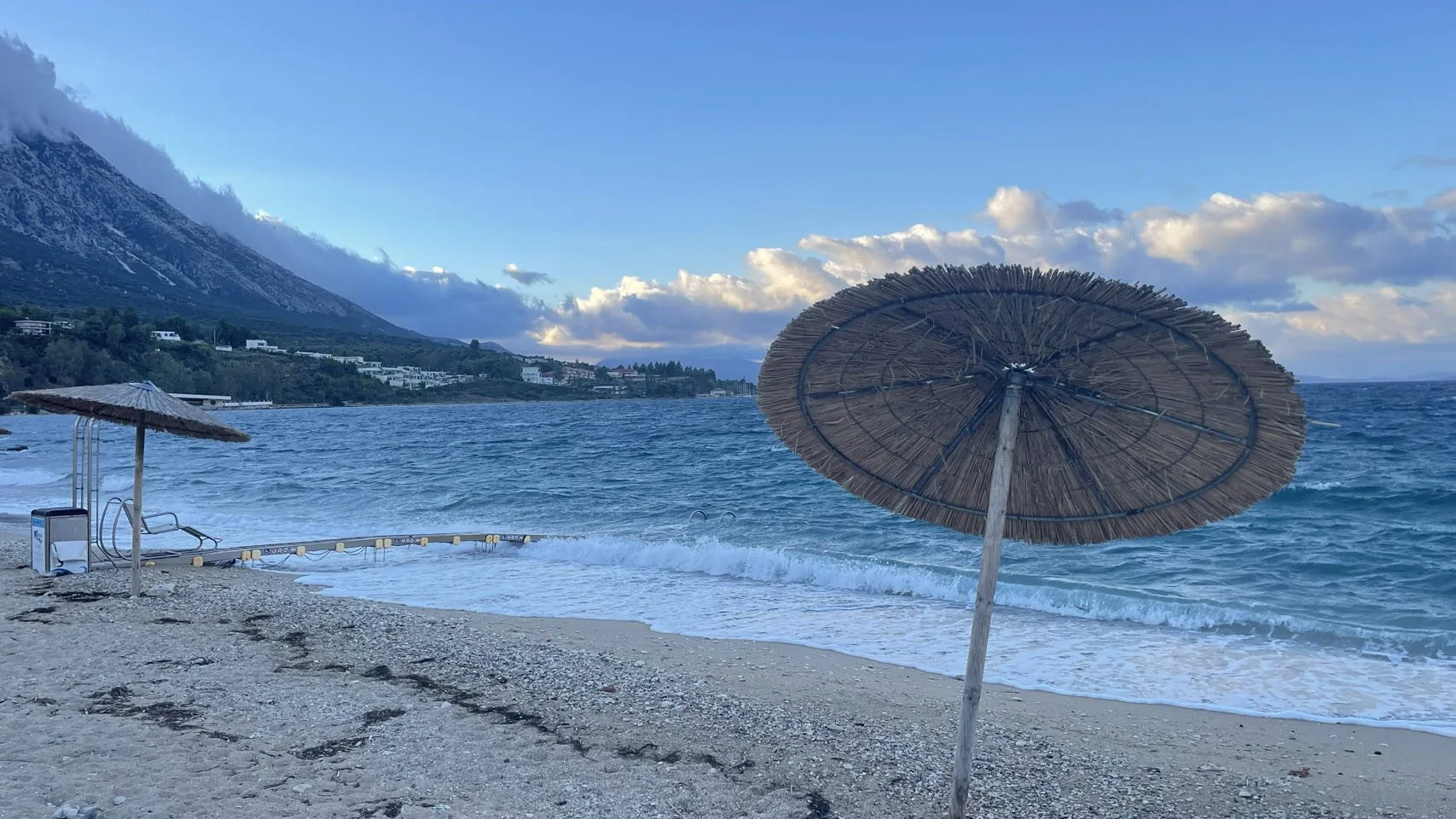 Een strand met twee parasols, één die rechtop staat en een grote die vrijwel volledig plat ligt, met zee en bergen op de achtergrond onder een bewolkte lucht.