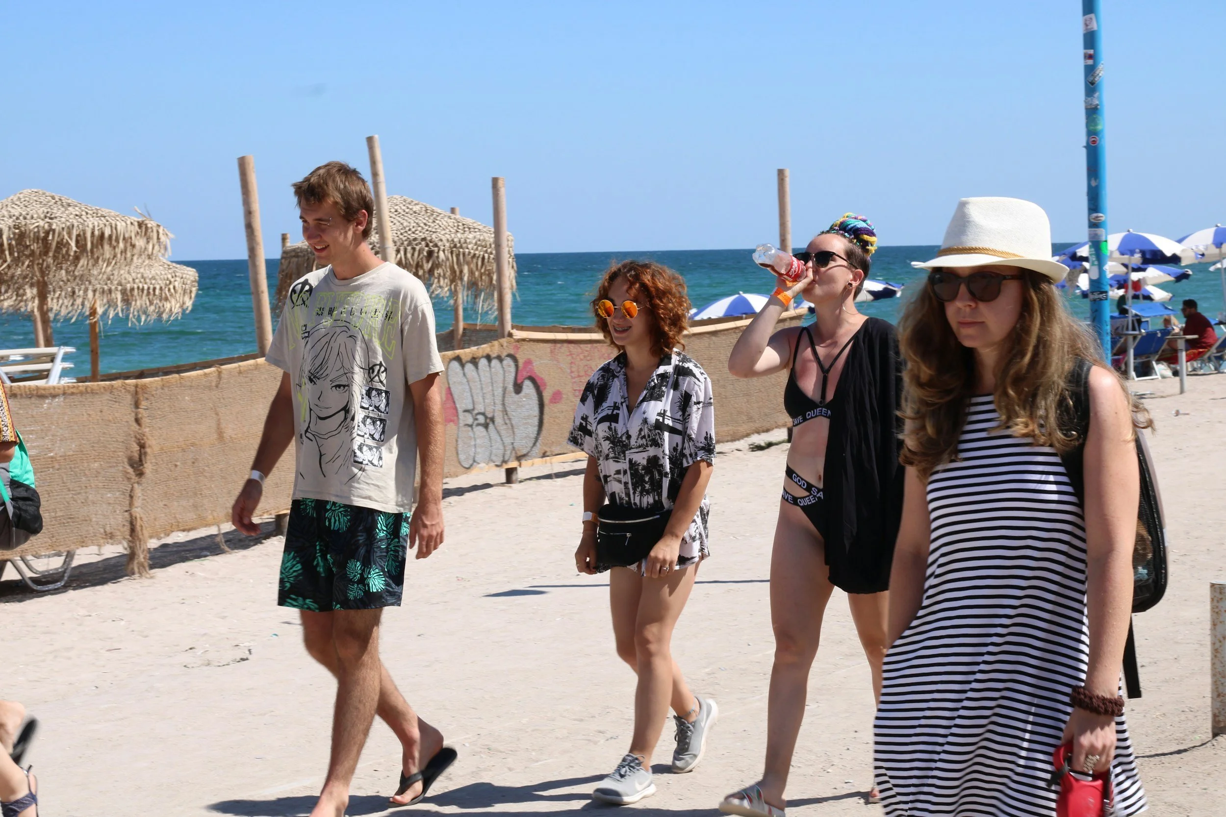 Vijf mensen lopen op het strand, allemaal dragen zomerkleding en zonnebrillen, met de zee op de achtergrond en parasols.