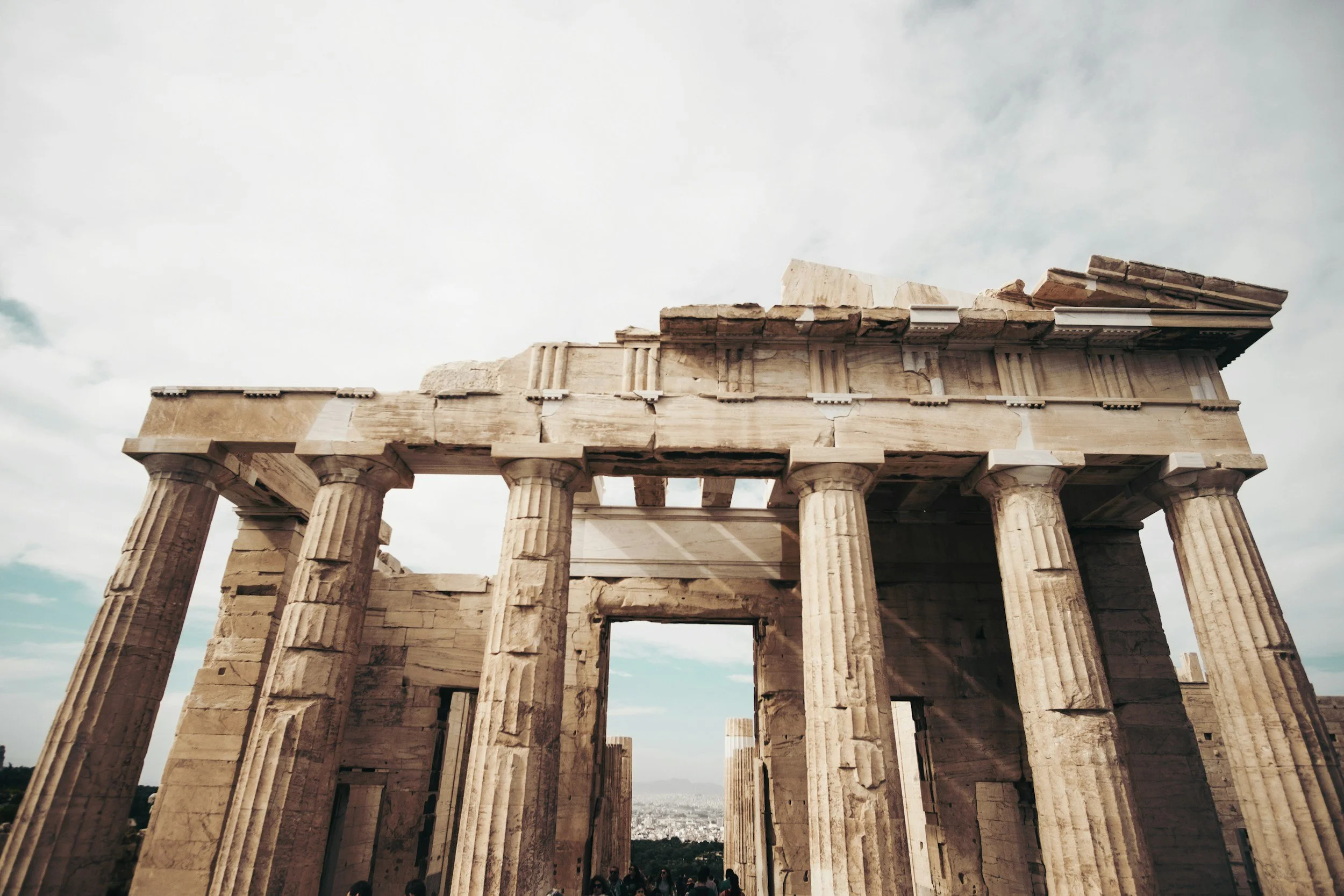 De ruïnes van het oude Griekse Parthenon-tempel met zes grote zuilen, onder een bewolkte lucht, gesproken door toeristen onderaan.