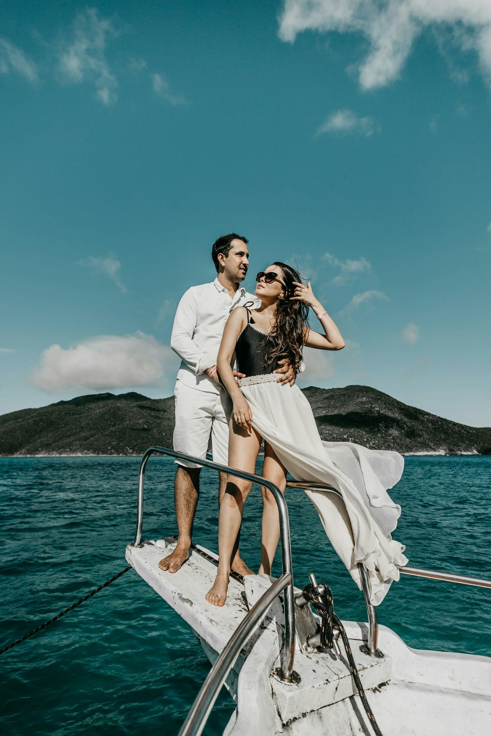 Een man en vrouw staan op het vooronder van een boot op het water, met een berg en een heldere blauwe lucht op de achtergrond.