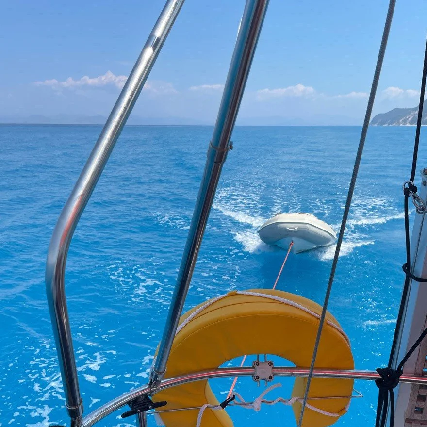 A boat met een gekromde gele life float en een witte opblaasbare waterdriehoek trekt een andere opblaasbare waterdriehoek door de oceaan, met op de achtergrond een eiland en een blauwe hemel.