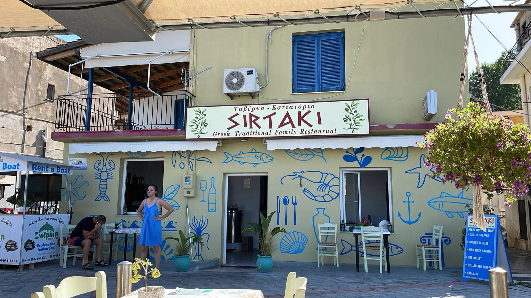 Een Grieks restaurant genaamd Sirtaki met blauwe visdecoraties op de gele gevel, buitenzitplaatsen en een menu bord met visgerechten. Mensen zitten en staan voor het restaurant.