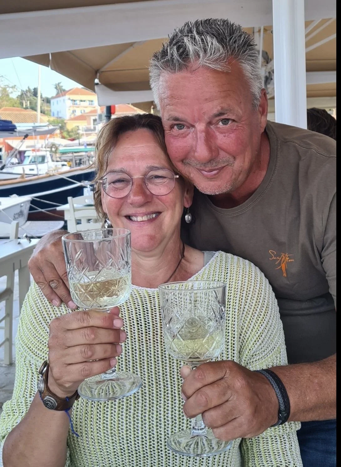 Twee mensen vieren met glazen wijn in een haven met boten op de achtergrond.
