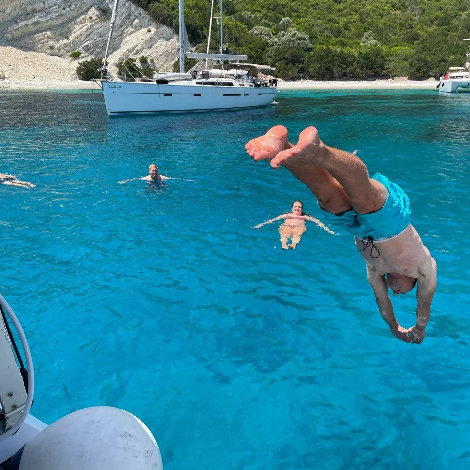 Persoon die achteroverkop over het water doet, andere mensen zwemmen in het water, boten liggen voor de kust, met groen bos en rotsen op de achtergrond.