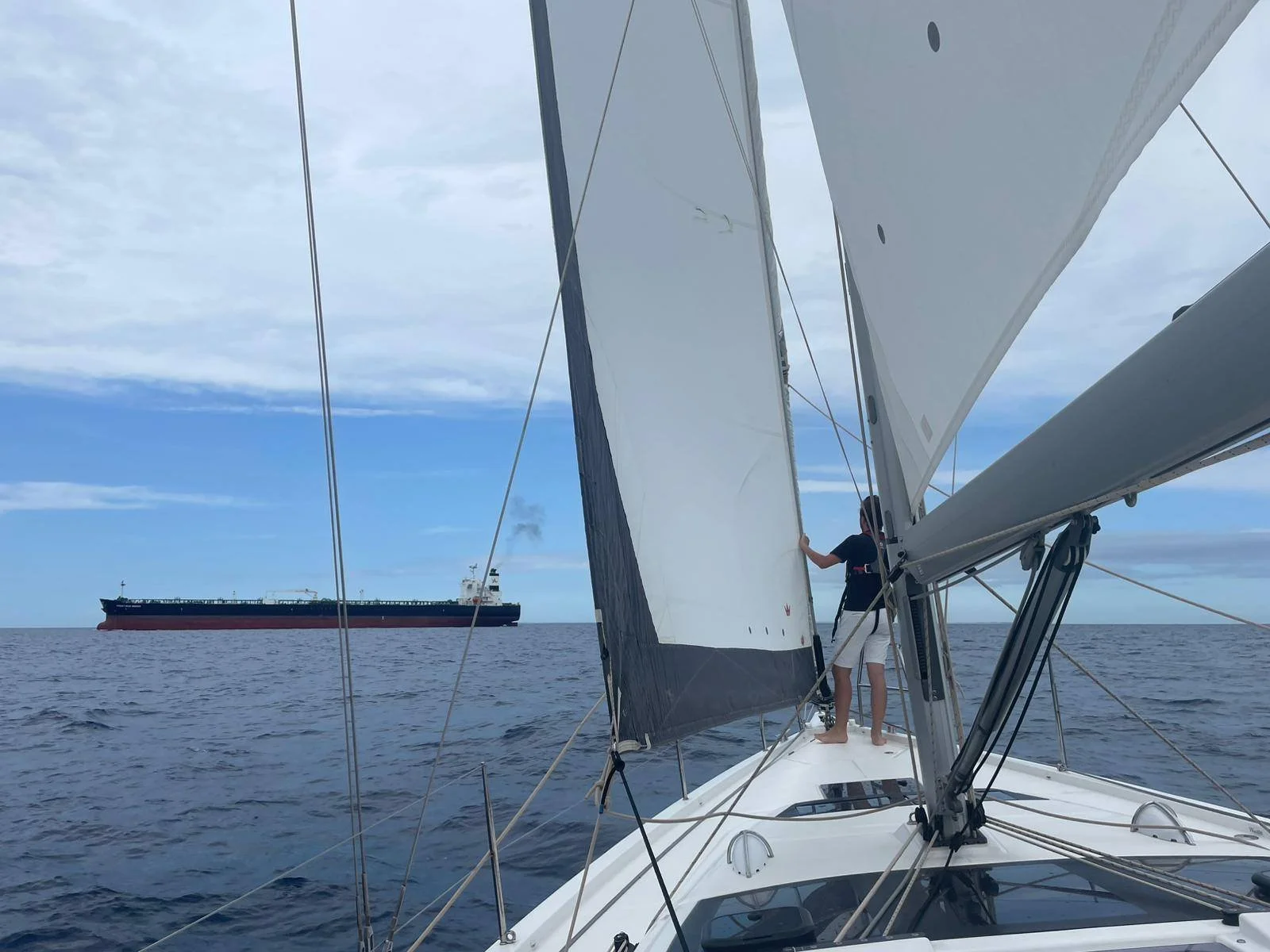 Een persoon staat op de boot met een bemanning die de zeilen bedient, een grote scheep vaart voorbij in de achtergrond onder een bewolkte hemel.