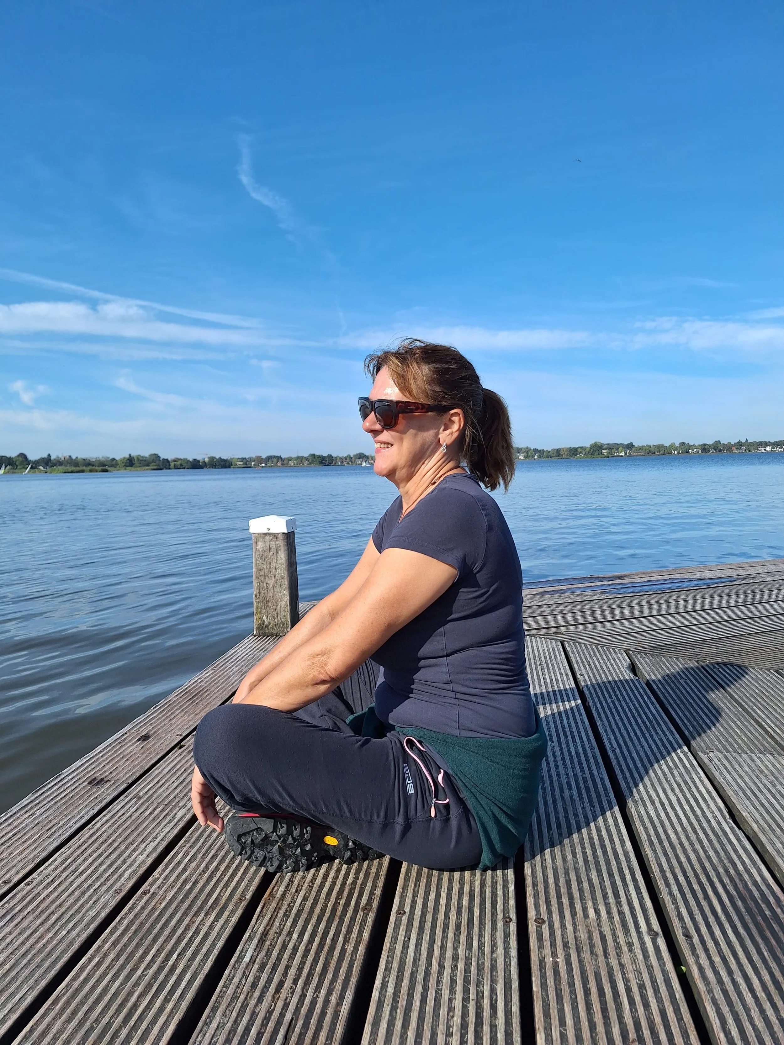 Vrouw die op een houten pier zit, met een waterpartij, een blauwe lucht en horizon op de achtergrond.