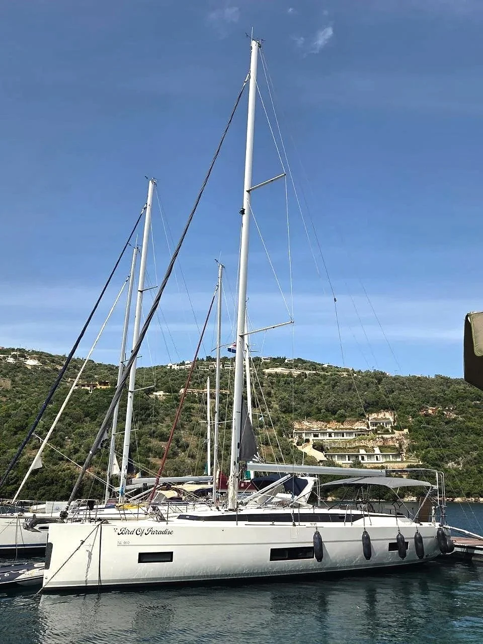 Een zeilboot genaamd 'Sea of Paradise' ligt aangemeerd in het water met een groene heuvel op de achtergrond. De boot heeft meerdere masten en is vastgemaakt met touwen.