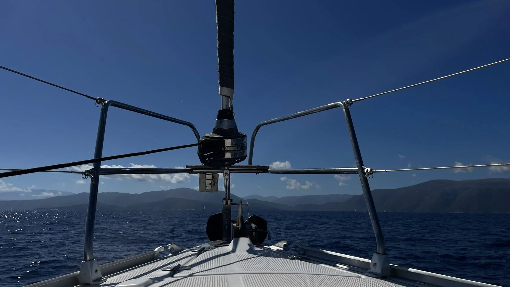 De voorkant van een zeilboot met stalen relingen, uitzicht op de oceaan en bergen op de achtergrond, onder een heldere blauwe hemel.