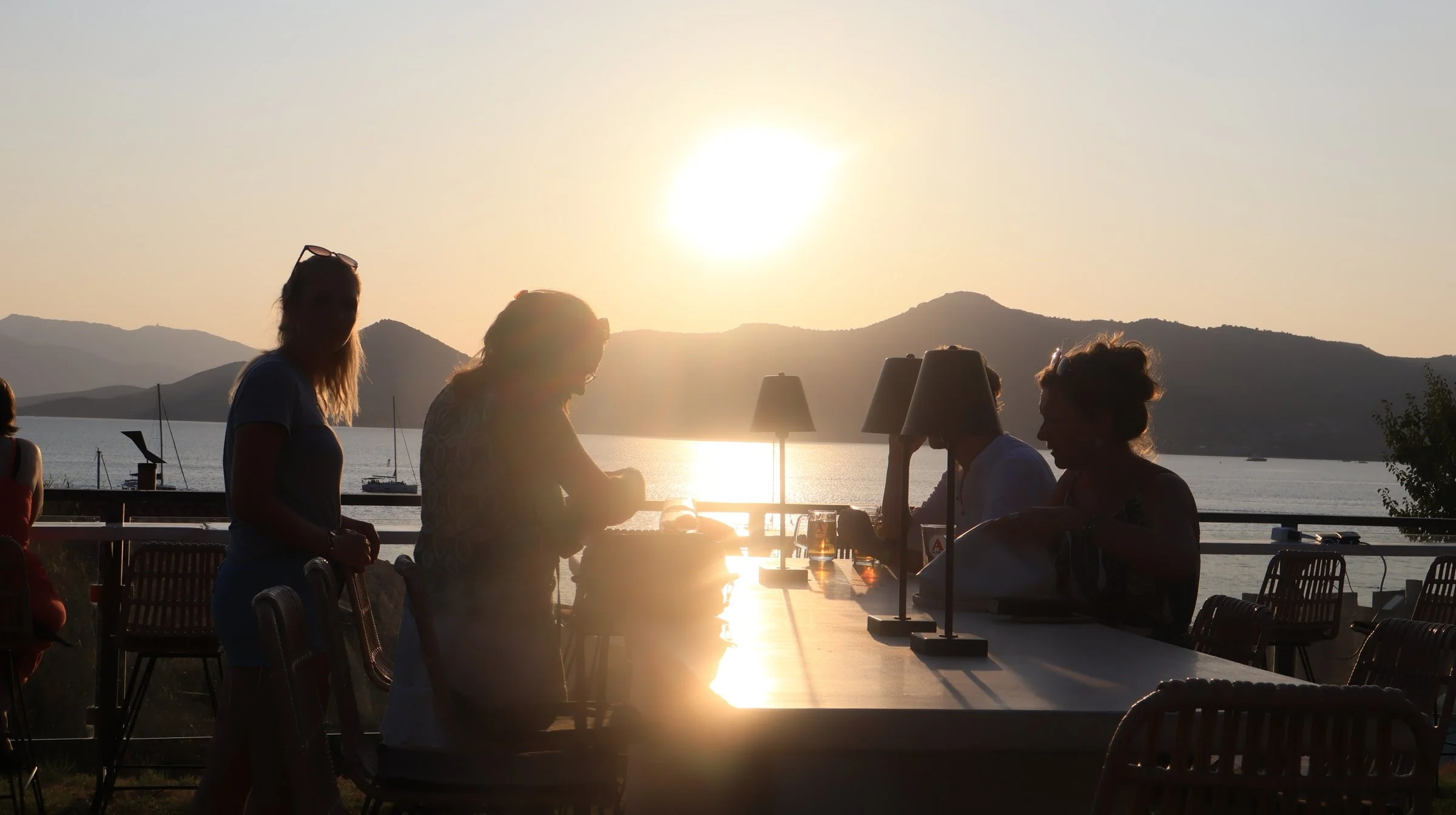 Mensen zitten bij een restaurant aan een tafel terwijl de zon ondergaat boven een meer en bergen.