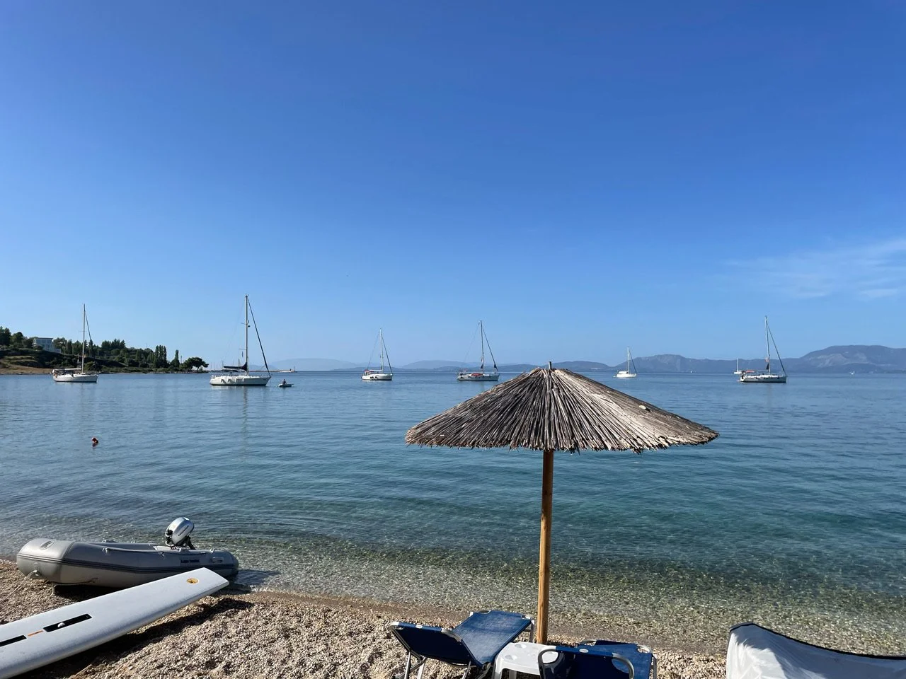 Een rustig meer met boten en een strand met een rieten parasol en ligstoelen in de voorgrond.