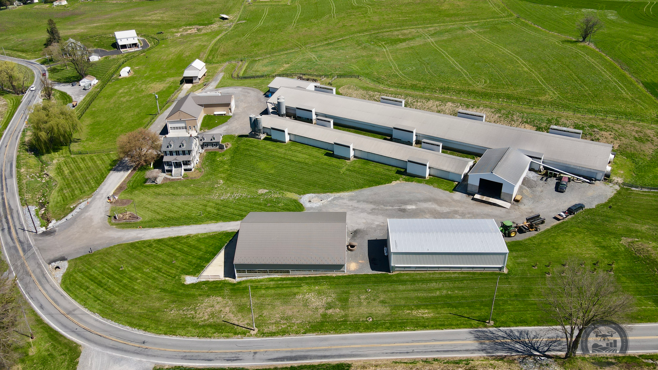 Aerial view of a rural property with a farmhouse, large barn, and several smaller outbuildings on green lawns, surrounded by fields and a curved road.