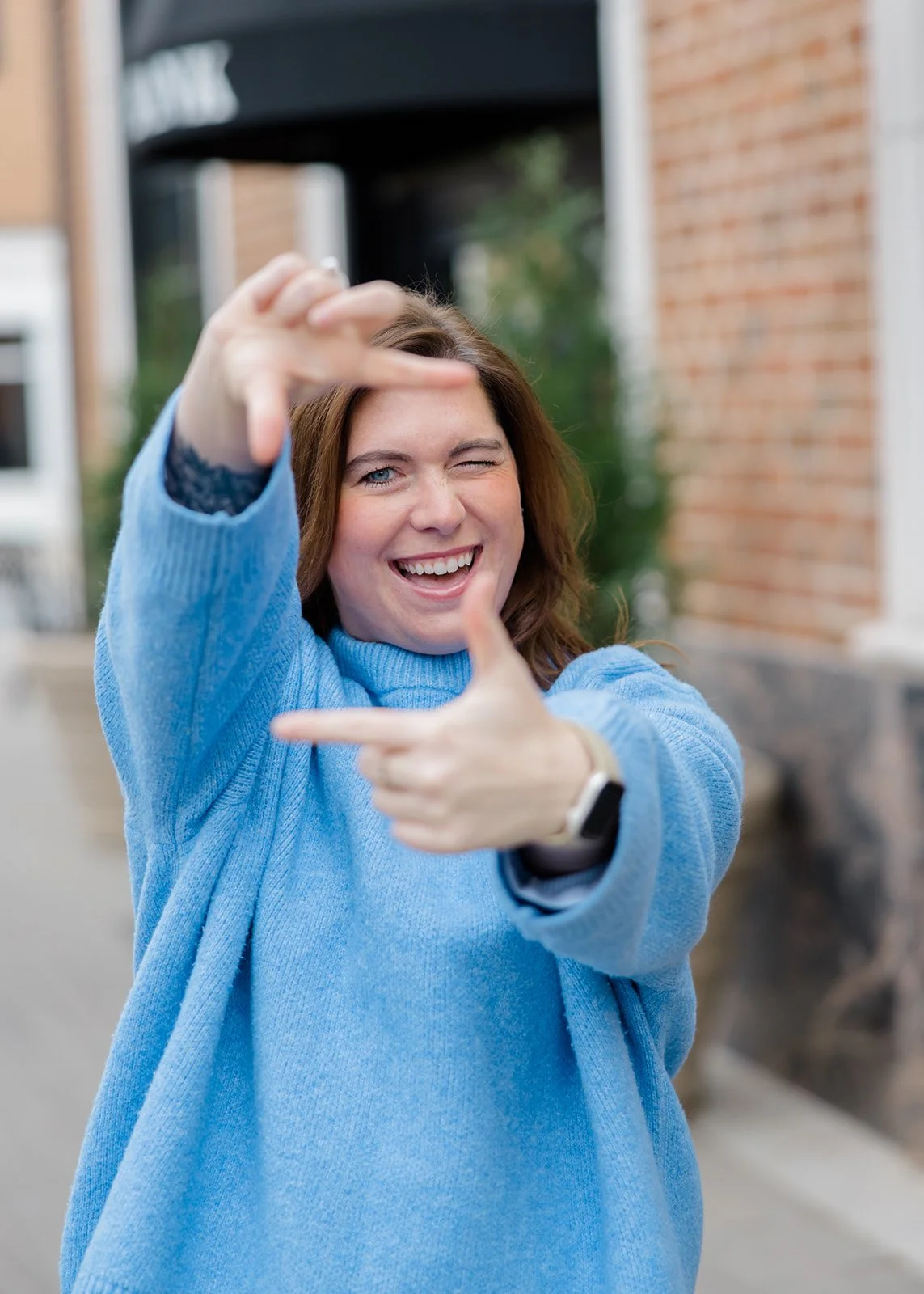A woman with long brown hair, wearing a blue sweater, winking and smiling while framing a rectangle with her hands.