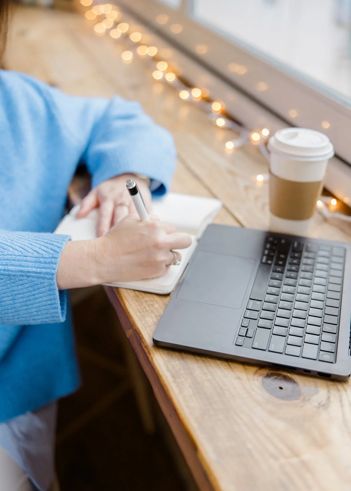 Person in a blue sweater writing in a notebook next to a laptop on a wooden table, with a coffee cup and string lights in the background.