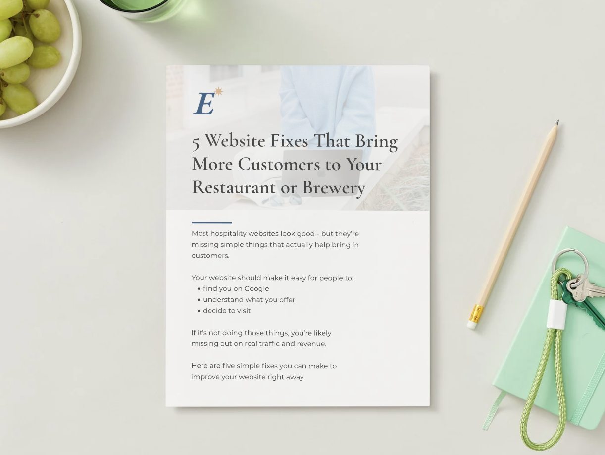 A printed article titled "5 Website Fixes That Bring More Customers to Your Restaurant or Brewery," placed on a light-colored surface next to a green notebook with keys and a stethoscope, with a bowl of green grapes and a drink nearby.