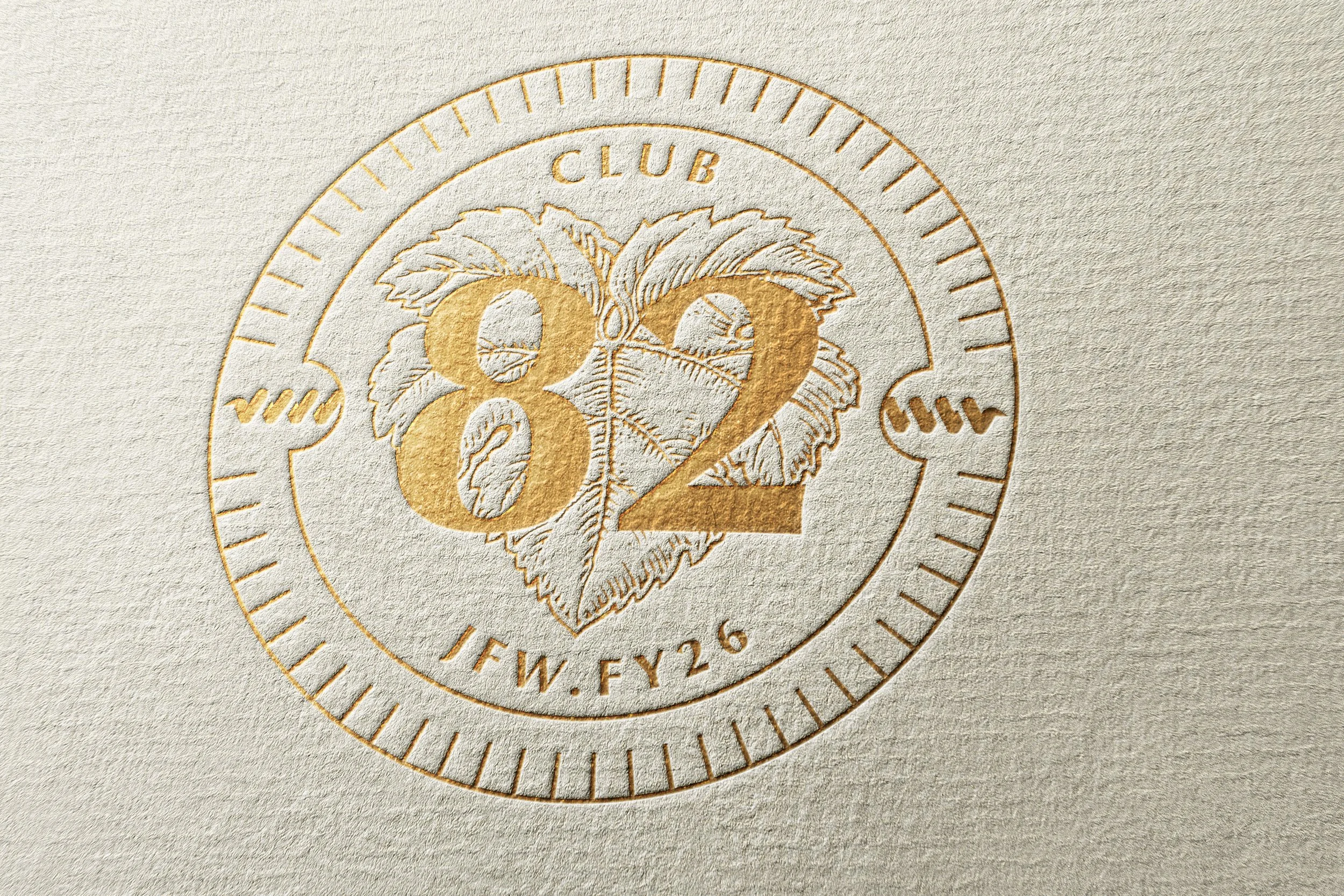 Embossed emblem with the number 82 in gold, featuring a leaf illustration, surrounded by the words 'Club' and 'IFW.FY26' on textured beige background.