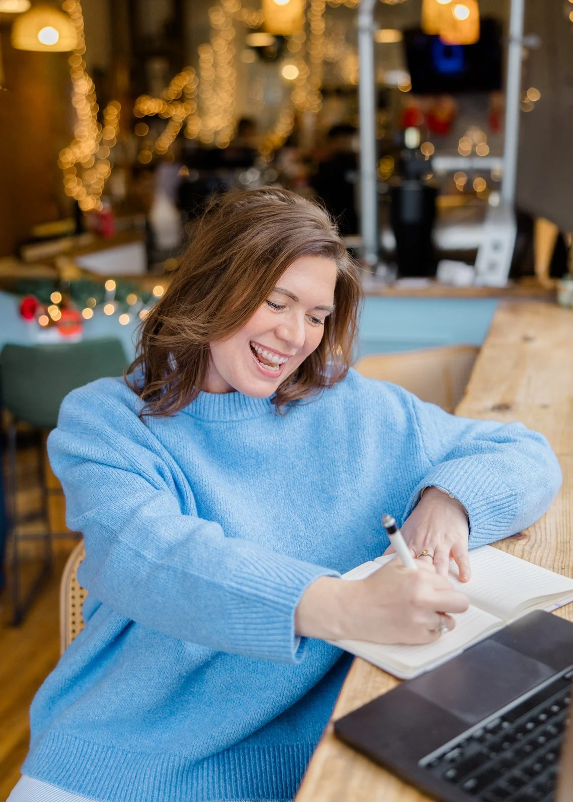 A woman in a blue sweater writing in a notebook at a wooden table in a lively cafe decorated with string lights.