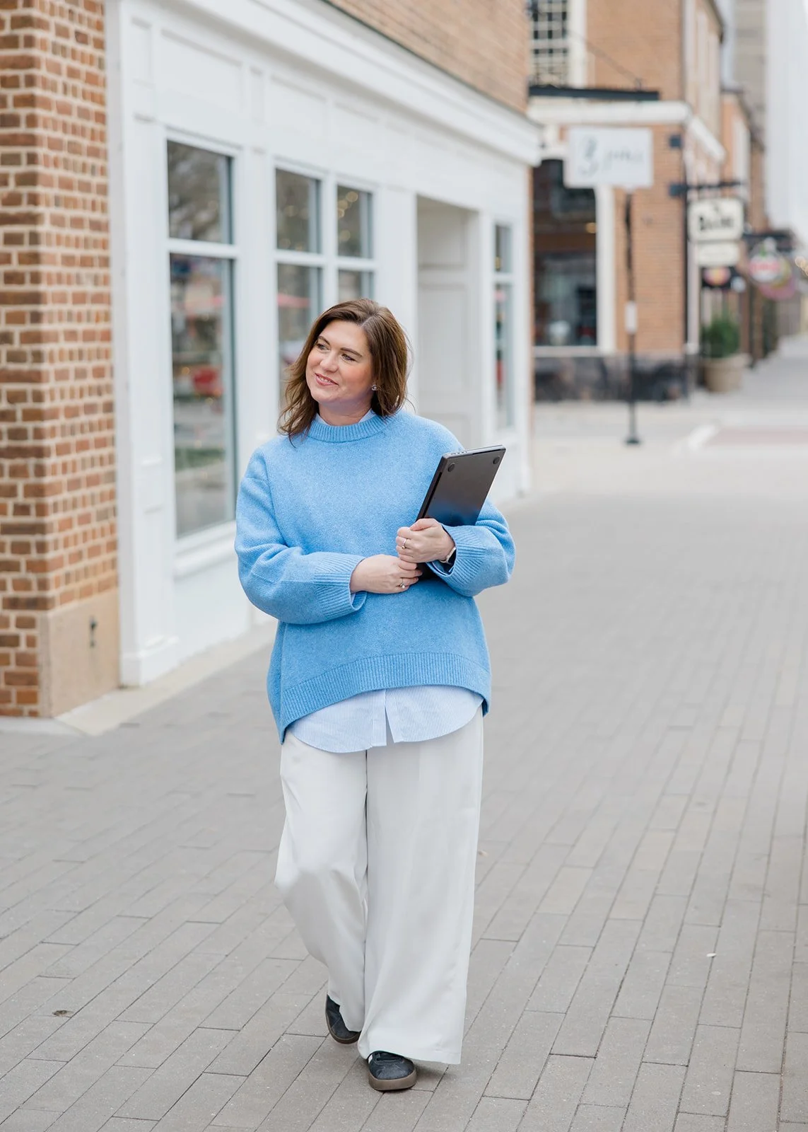A woman walking on a city sidewalk holding a tablet device, wearing a light blue sweater, white shirt, and white pants.