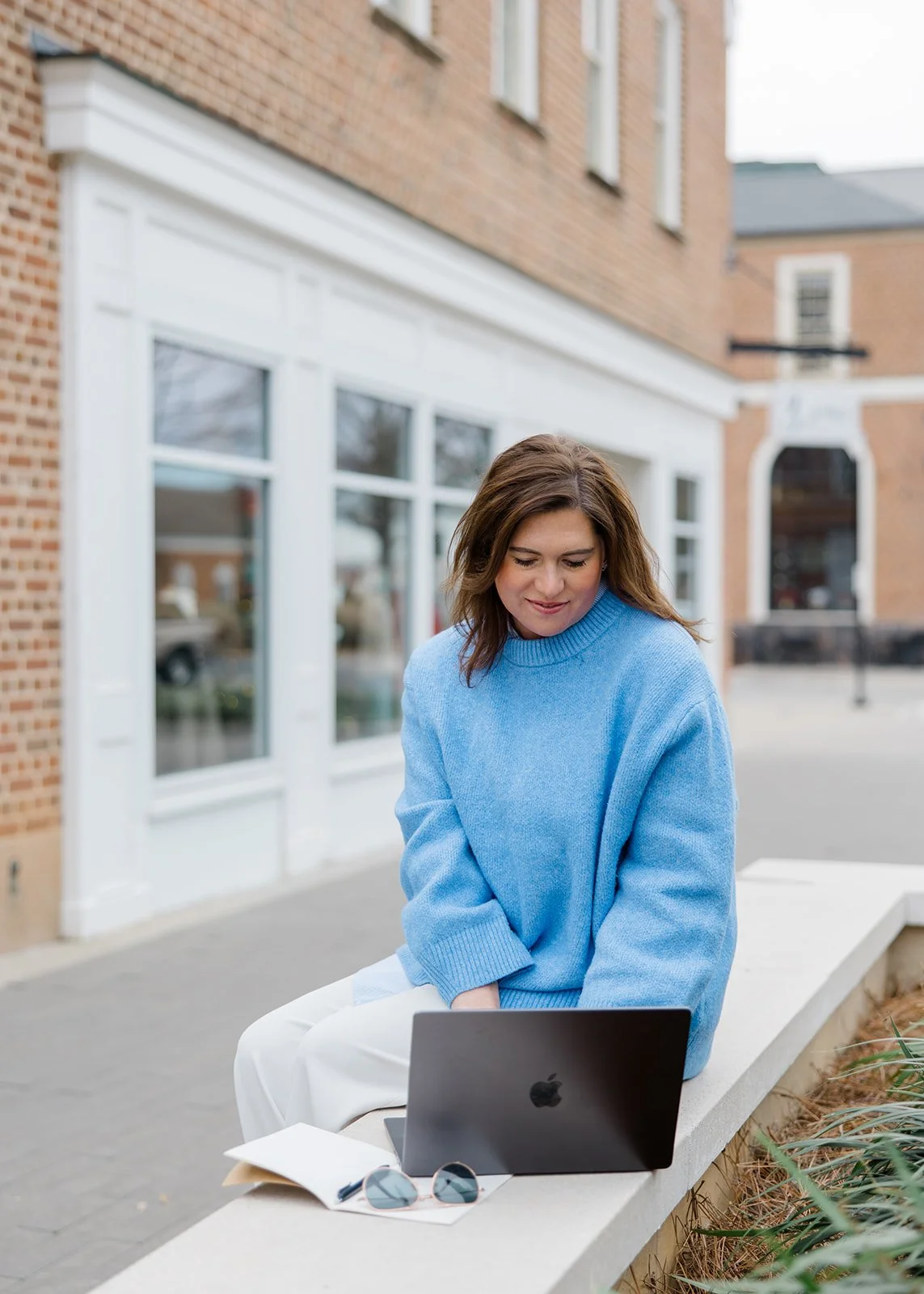 Woman sitting on a concrete bench outdoors using a laptop with a building with large windows behind her.