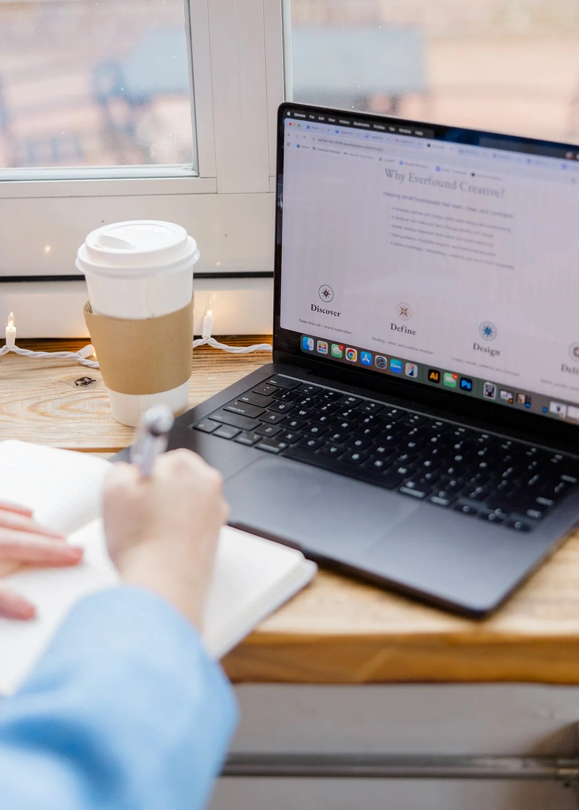 A person writing in a notebook at a wooden table, with an open laptop displaying a website, a coffee cup with a cardboard sleeve, and a window in the background.
